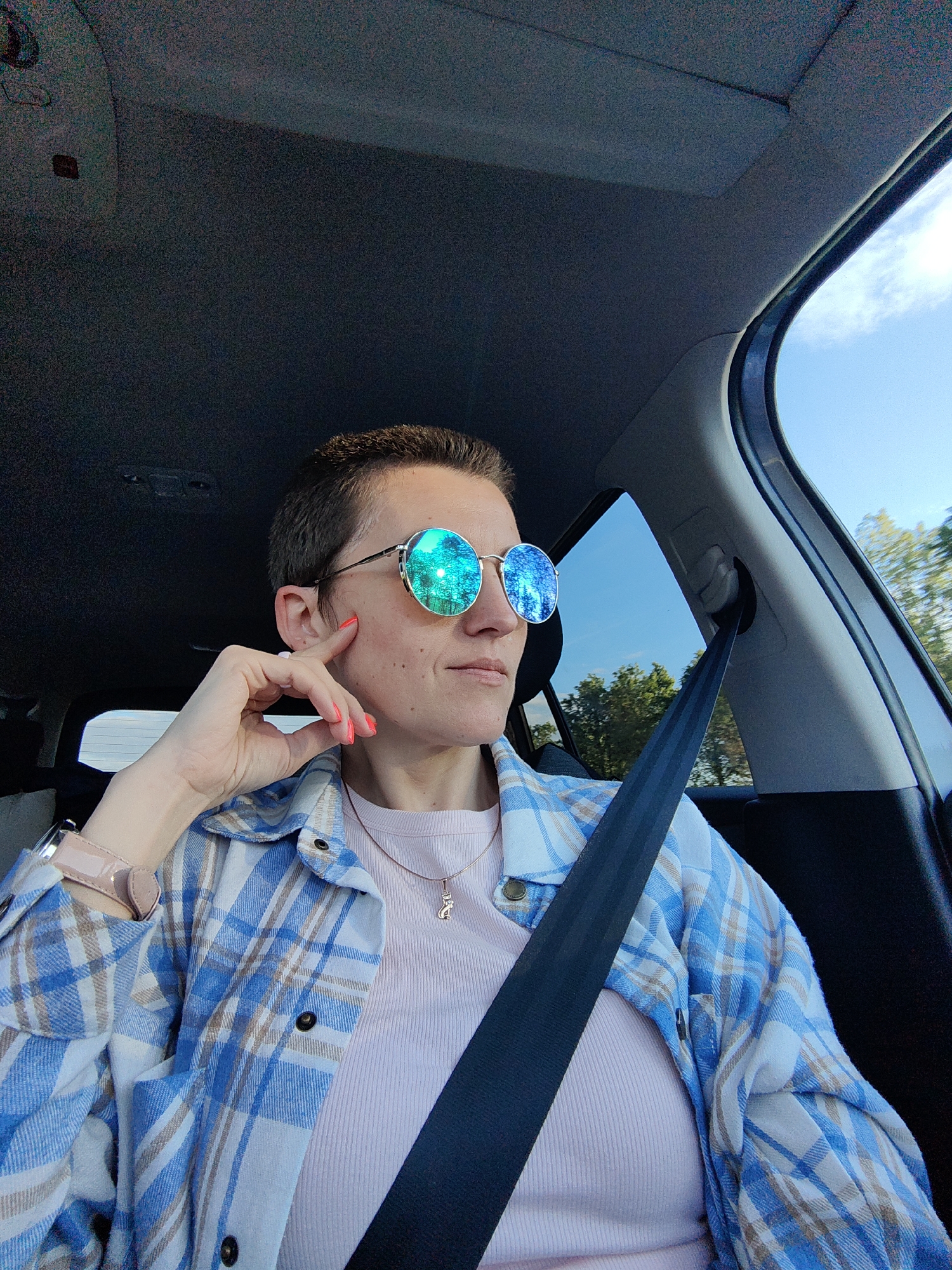 A woman with short, dark hair is sitting in the passenger seat of a car, wearing round sunglasses with mirrored lenses that reflect a green forest. She has a light pink ribbed shirt on and an unbuttoned blue and white plaid shirt over it. A delicate necklace with a cat pendant is visible. Her right hand is resting on her cheek, with bright coral-colored nail polish on her fingers. A car seatbelt is visible across her chest. The background outside the car window shows a clear blue sky and trees, suggesting it is daytime. The interior of the car is dark, with the headliner and a portion of the car's pillar visible. The scene is likely a snapshot taken during a car journey in the Фаніпальскі сельскі Савет, Belarus.
