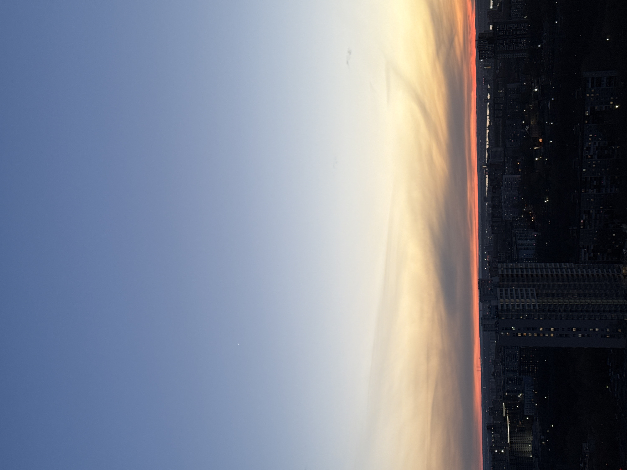 This image presents a panoramic view of a city at twilight, capturing the transition between day and night from a high vantage point.
The sky dominates the upper two-thirds of the frame, displaying a striking gradient of colors. On the left, it is a clear, deep blue, gradually lightening towards the center and right. Here, the sky transitions into soft hues of yellow and orange, culminating in a vibrant, thin band of reddish-pink along the horizon line. This intense coloration suggests either a recent sunset or an impending sunrise, with the sun just below the visible horizon. A small, bright white dot, likely a prominent star or planet like Venus, is distinctly visible in the upper-left quadrant of the blue sky.
The cityscape occupies the lower third of the image, appearing as a dark silhouette against the illuminated sky. Numerous buildings of varying heights are discernible, many with illuminated windows, indicating residential or commercial activity within the urban environment. Streetlights and other artificial lights also dot the dark urban fabric, creating a pattern of glowing points. Along the distant horizon line, faint outlines of what appear to be industrial structures or tall chimneys can be observed rising slightly above the general building mass.
No people or explicit activities are visible, emphasizing the quiet, vast expanse of the twilight hour over the city. The weather is clear, with minimal cloud cover, allowing for an unobstructed view of the atmospheric effects. No visible text is present in the image. The scene captures a serene and expansive moment over a bustling urban center.