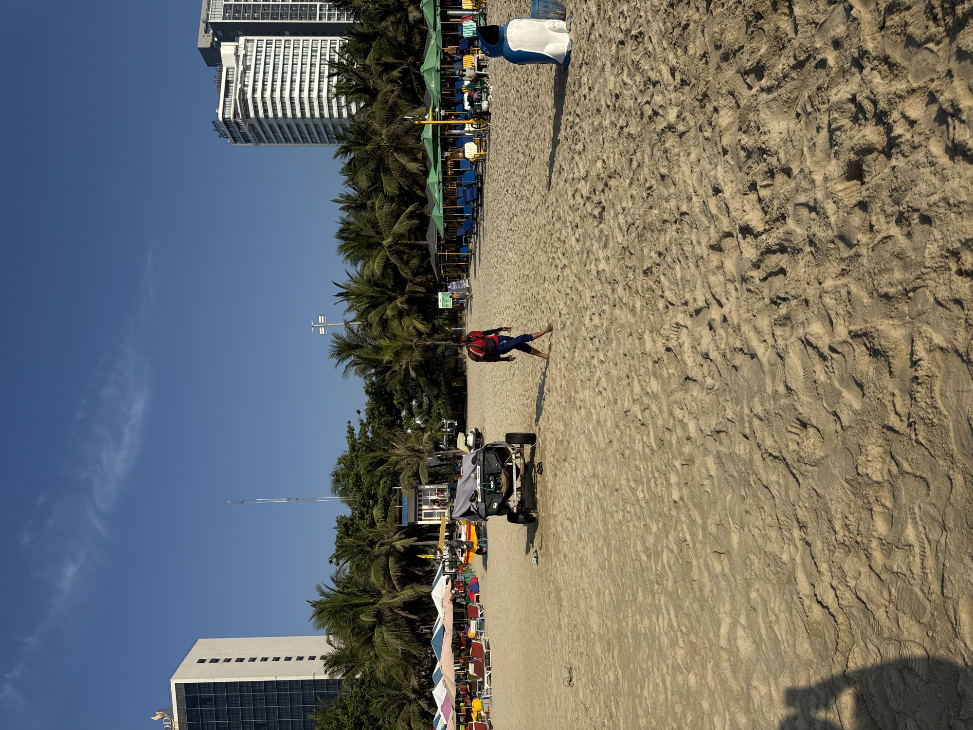 The image captures a beach scene in Đà Nẵng, Vietnam. The scene is split between a sandy beach occupying the lower right and a clear blue sky in the upper left. Along the beach, rows of palm trees delineate the boundary between the sand and the urban development. Several high-rise buildings are visible in the background, suggesting a city beach setting. Various objects are scattered along the beach, including beach umbrellas, chairs, and what appears to be a lifeguard vehicle. A person is seen walking barefoot on the sand. The beach shows signs of activity, with numerous footprints visible in the sand.