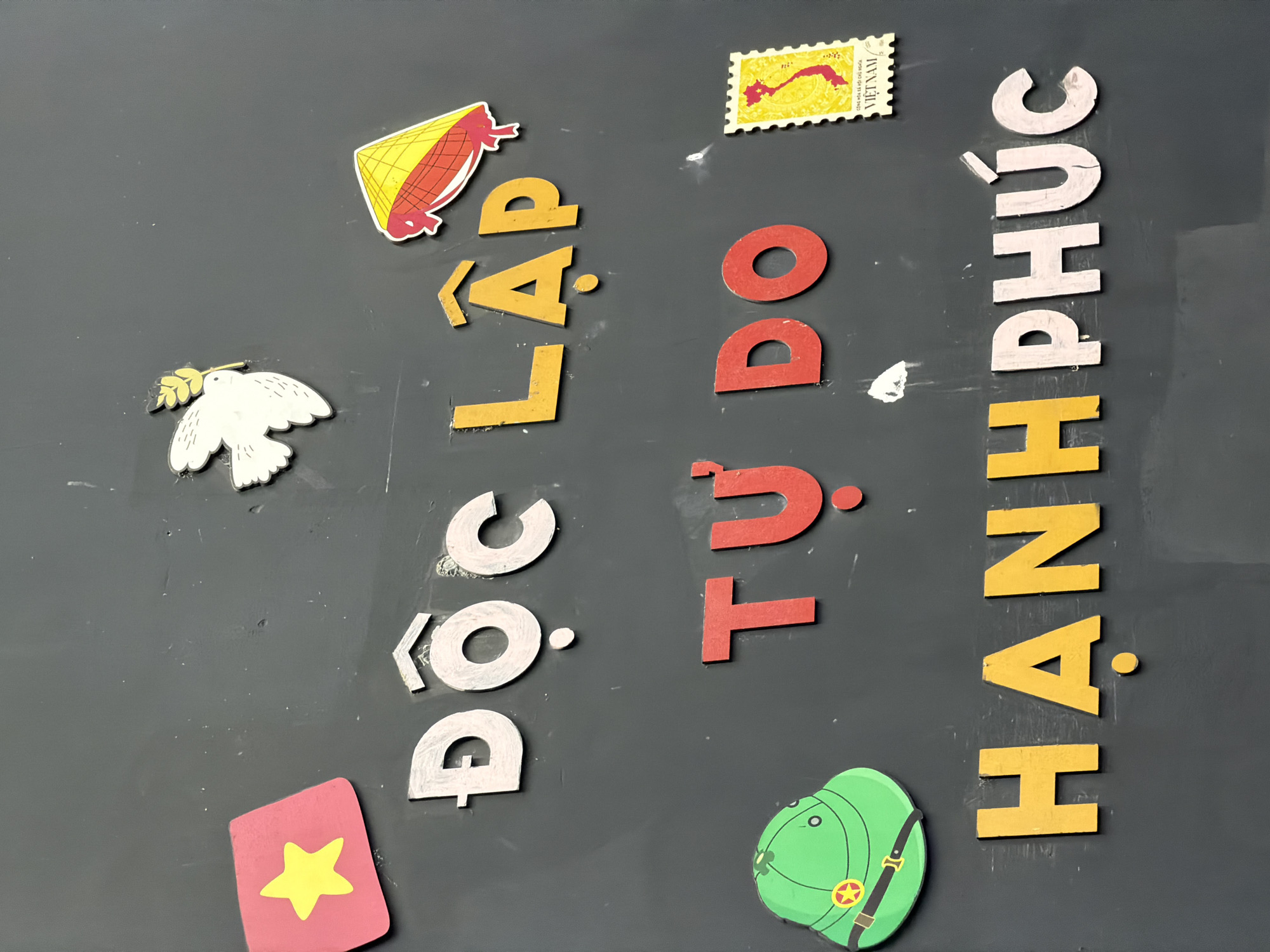 This image displays a decorative arrangement on a dark, possibly magnetic, surface, likely a board. The primary subjects are magnetic letters forming Vietnamese words and various symbolic cut-outs.
The most prominent feature is the text, which reads "Độc lập, Tự do, Hạnh phúc" when read vertically from left to right as intended. This translates to "Independence, Freedom, Happiness," a well-known national motto of Vietnam. The letters are made of cut-out material, with "Độc lập" primarily in white and yellow, "Tự do" in red, and "Hạnh phúc" in yellow and white.
Several culturally significant items are scattered around the text. On the left, a white dove holding an olive branch symbolizes peace, positioned above a red, rectangular cut-out featuring a yellow five-pointed star, reminiscent of the Vietnamese flag. On the right, at the top, there is a traditional Vietnamese conical hat (Nón Lá) in yellow and red. Below it, a yellow postage stamp clearly visible with "VIỆT NAM" written on it. Towards the bottom right, a green military-style helmet with a red and yellow star emblem is displayed, often associated with the People's Army of Vietnam.
The dark surface shows minor scuff marks and white specks, indicating use. The items appear to be made of sturdy paper, foam, or thin wood, possibly with magnetic backing. The arrangement suggests a patriotic or educational display, celebrating Vietnamese national values and culture. Given the context, this display is located in Đà Nẵng, Vietnam.