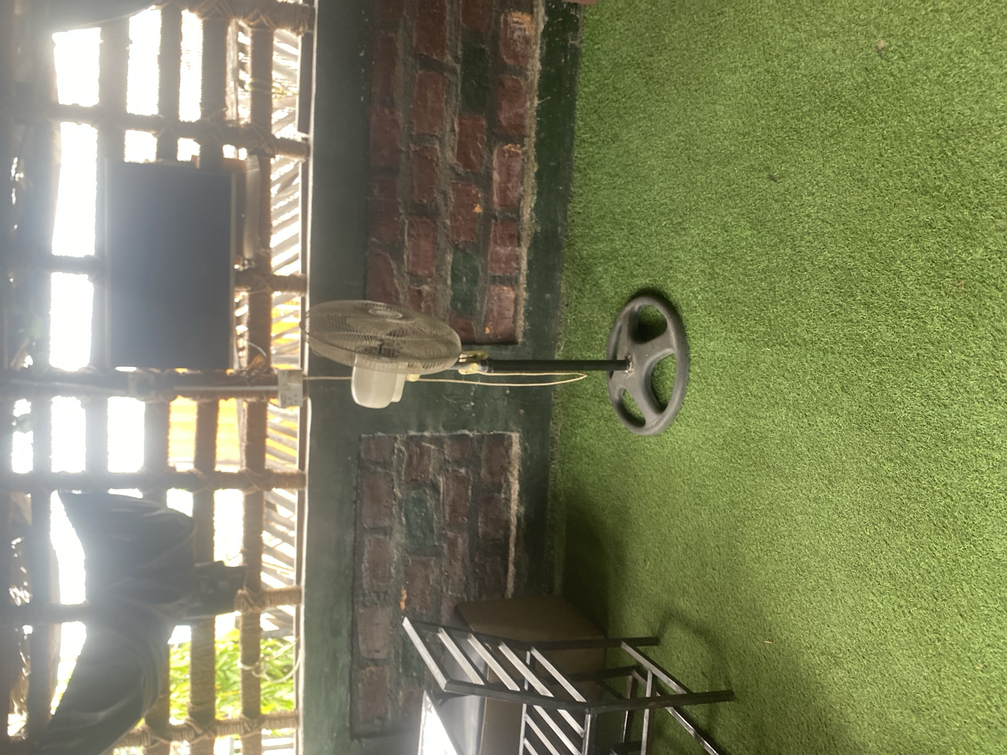 The image depicts an indoor space with a rustic aesthetic, possibly a gym or a recreational area in Jalingo, Nigeria. The floor is covered with green artificial turf, resembling grass. To the left, a large window or opening with a wooden lattice structure allows bright sunlight to stream in, creating a strong backlighting effect. A dark, rectangular object, possibly a television or a screen, is mounted within this structure.
In the center of the image, a white pedestal fan is positioned on the turf. It is oriented towards the right, with its blades visible. Further to the right, a black weight plate, typically used in weightlifting, rests on the artificial grass. Behind the fan, a wall constructed of rough, reddish-brown bricks, partially covered with green paint, serves as a backdrop. A black metal structure, resembling a rack or a set of chairs, is partially visible in the lower right portion of the frame. The overall impression is one of an unfinished or casually decorated space. The lighting suggests it might be daytime. There is no visible text in the image.