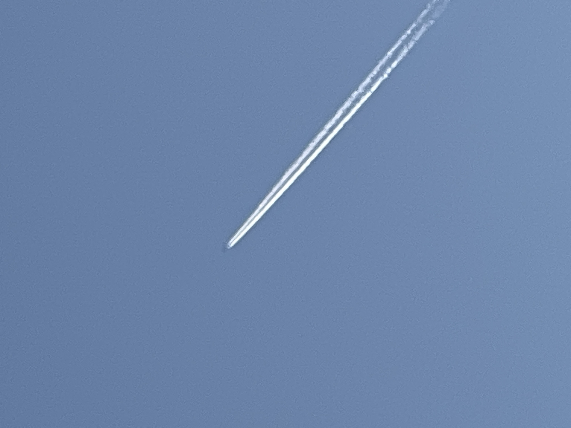 The image depicts a contrail in a clear, blue sky. There are no people, distinct objects, or discernible actions in the scene. The contrail is the only subject, appearing as a white line tapering off into the distance. The environment is a clear, unobstructed sky. There is no evidence of location in Chaikovsky, Russia, or any other specific place.