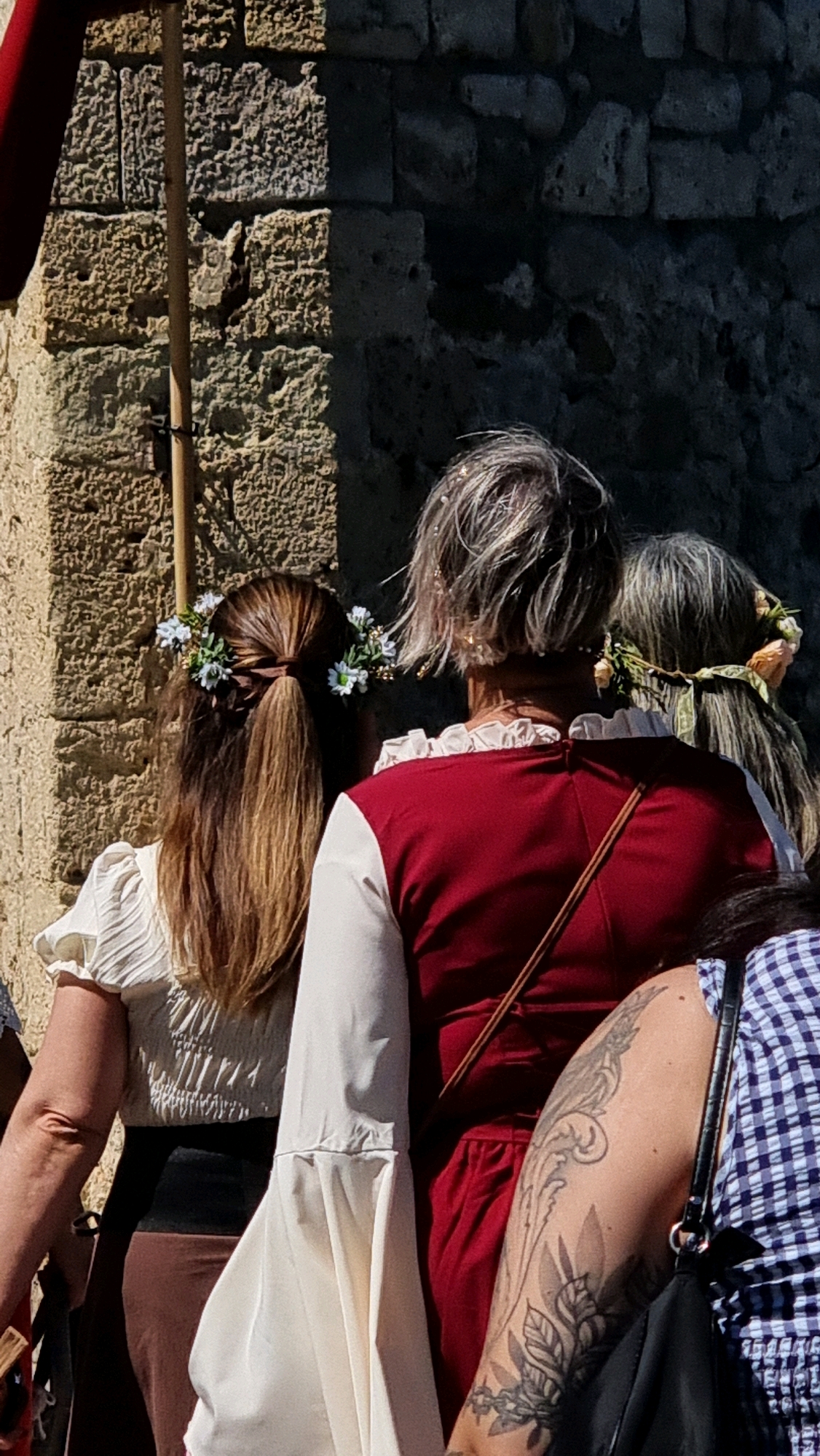 The image shows the backs of several people dressed in what appears to be historical or Renaissance-era attire. The primary focus is on the individuals in the foreground, who are walking alongside a stone wall.
In the foreground, a person with long, brown hair adorned with a floral crown is seen from behind. They are wearing a cream-colored top with puffed sleeves and a dark skirt. To their right, another person is visible, wearing a deep red garment with a cream-colored ruffled collar and long cream sleeves. This person has short, grey hair, also adorned with some glitter or small ornaments. Further to the right, a third person is partially visible, with grey hair also decorated with a floral crown. The arm of another individual on the far right, sporting a tattoo, is also in view, wearing a blue and white checkered garment.
The background consists of a rough, textured stone wall, typical of old European architecture. The lighting suggests a sunny day. The context provided indicates that this scene is located in the city of Besalú, Spain, a town known for its well-preserved medieval architecture, which aligns with the visual elements in the image. The people appear to be participating in a historical reenactment or festival, given their clothing and the environment.