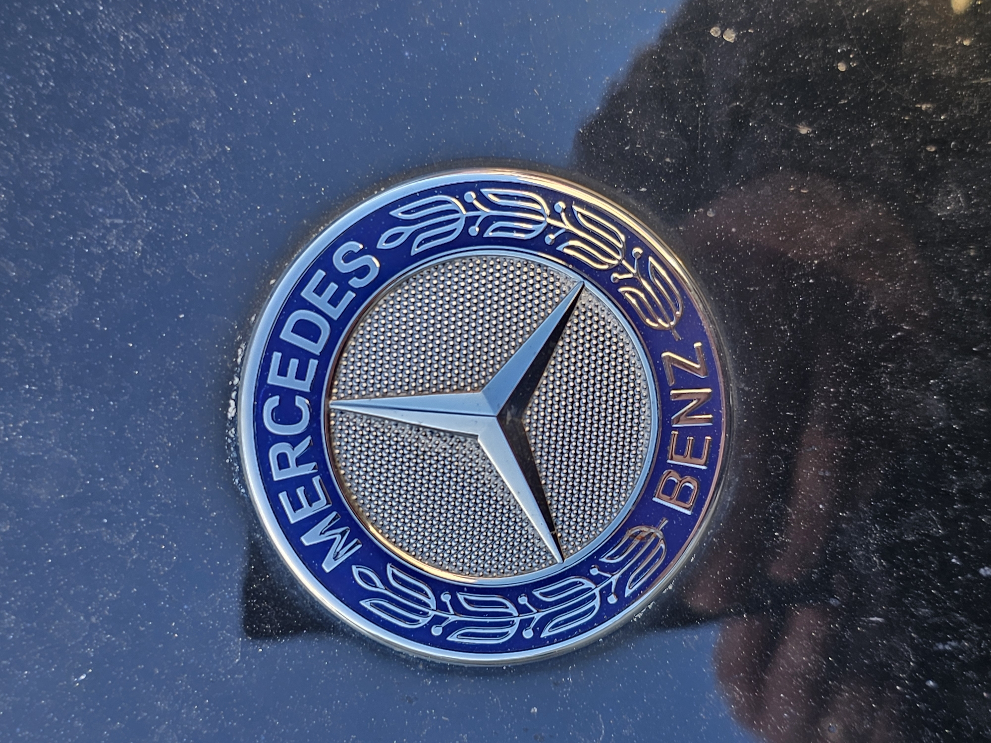 A close-up, slightly elevated perspective captures a Mercedes-Benz emblem mounted on a dark, glossy vehicle surface, likely a car hood or trunk. The circular emblem features a prominent silver, three-pointed star at its center, set against a textured, dimpled silver background. The star itself is reflective, showing highlights. Surrounding the central star, the outer ring of the emblem is dark blue, emblazoned with the silver text "MERCEDES" on the left and "BENZ" on the right. Stylized silver laurel wreath patterns adorn the blue ring between and around the text. The emblem appears clean and polished, contrasting sharply with the surrounding dark surface of the car, which is noticeably covered in a layer of fine dust and small particles. Reflections are visible on the car's surface, with a faint, distorted human-like shape, possibly the photographer's reflection, noticeable in the bottom right corner of the image. The lighting indicates an outdoor setting, possibly daylight, illuminating the emblem's metallic sheen. The scene is located in Holywell, United Kingdom.