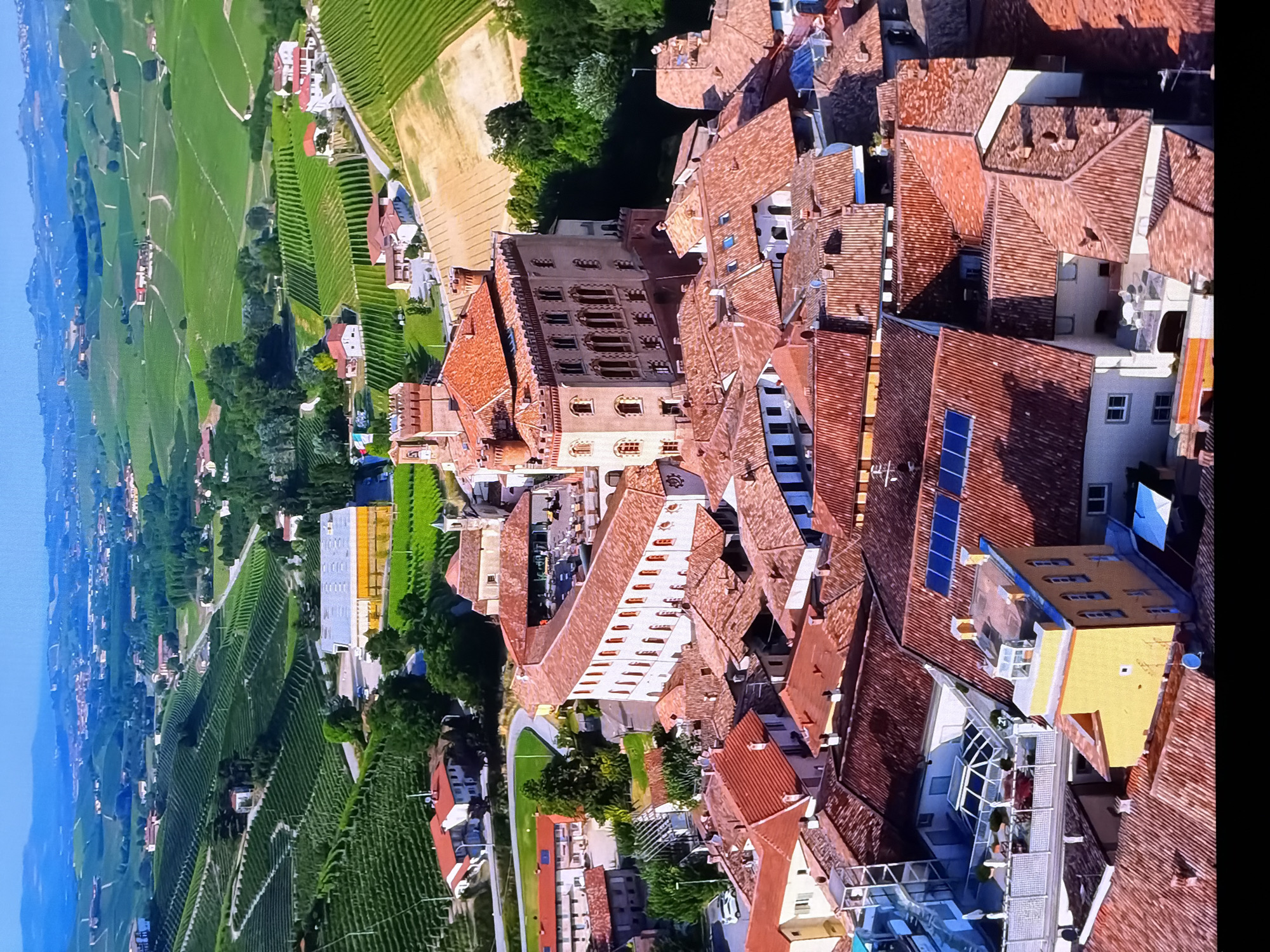 This is an aerial, wide-angle shot of a historic European town nestled amidst rolling green hills and vineyards. The scene is dominated by terracotta tiled rooftops, a large, imposing building with a red-tiled, multi-tiered roof and fortified towers, possibly a castle or large manor. This central structure is surrounded by a dense cluster of other buildings with similar red tiled roofs.
In the mid-ground, a large, modern building with a yellow facade and a flat roof is visible. Adjacent to it, a contemporary structure with a glass atrium and an outdoor seating area is present. The surrounding landscape is characterized by meticulously organized vineyards covering the hillsides, with some patches of bare earth and scattered trees. In the far distance, more rolling hills and a hazy blue sky are visible.
There are no people visible in the image. The scene depicts a peaceful, picturesque, and seemingly well-maintained urban and rural environment. The location is not Chirchik, Uzbekistan.