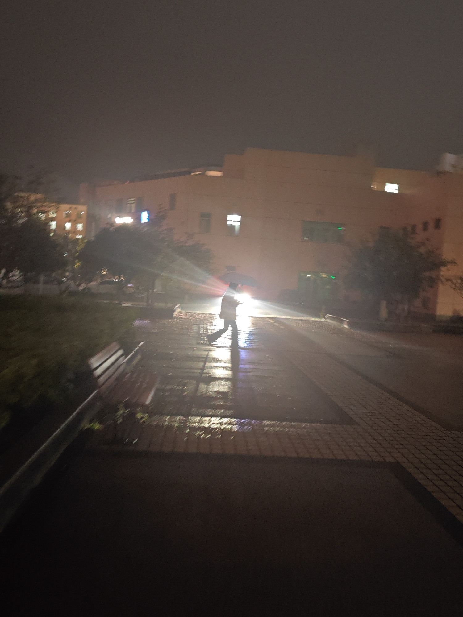 A night scene in Jiangning District, China, shows a wet brick pathway illuminated by light, likely from a vehicle, reflecting off the damp surface. A person is walking away from the light source, their form indistinct. Buildings are visible in the background, with lit windows. A bench is partially visible on the left, bordering a grassy area. Trees are present on both sides of the pathway. The overall impression is of a rainy night in an urban area.