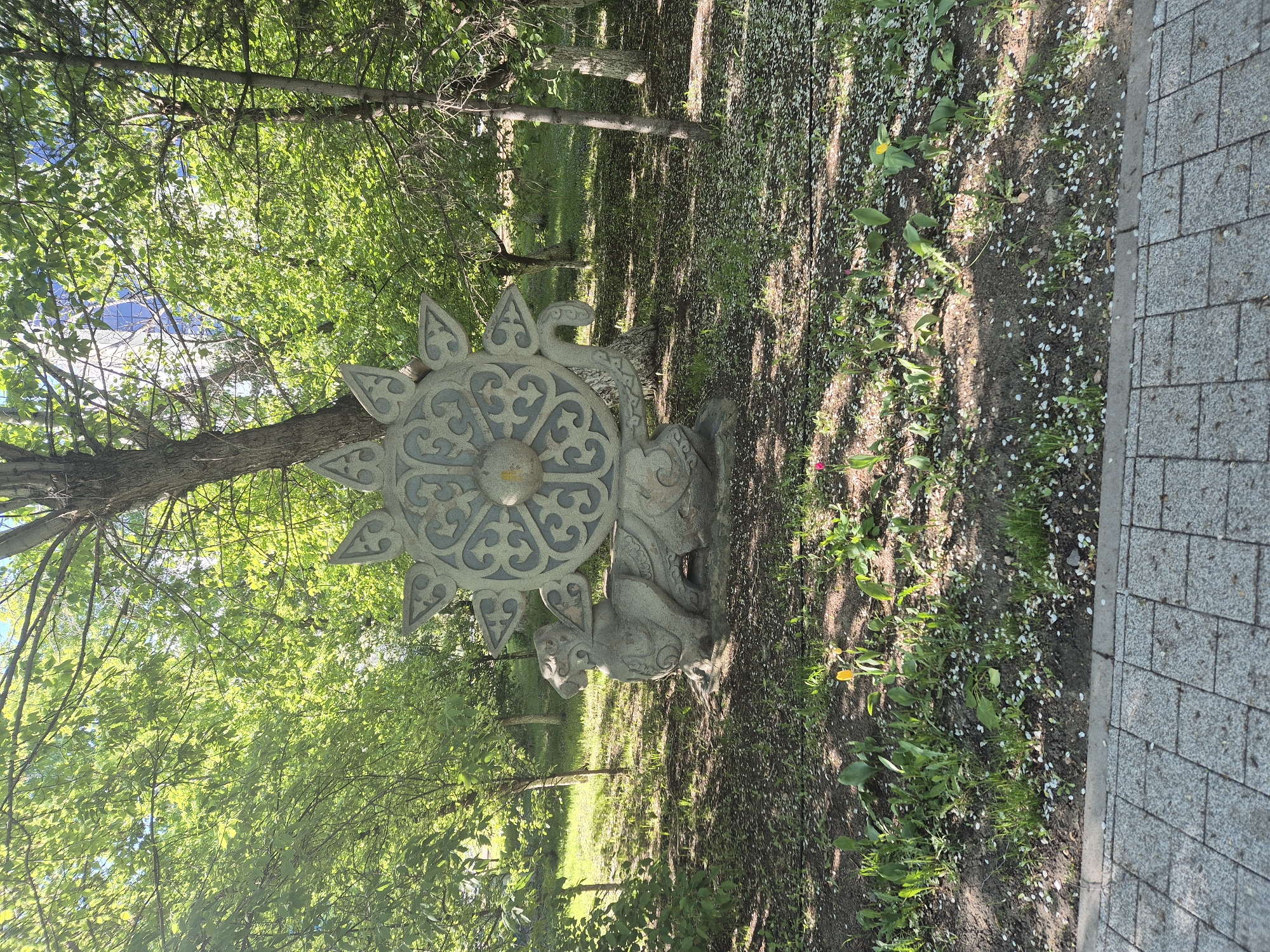A stone monument with a sun-like design is positioned in a green space near a paved path. The monument sits among trees, grass, and scattered white petals. The location appears to be a park in the city of Unknown, Unknown.
