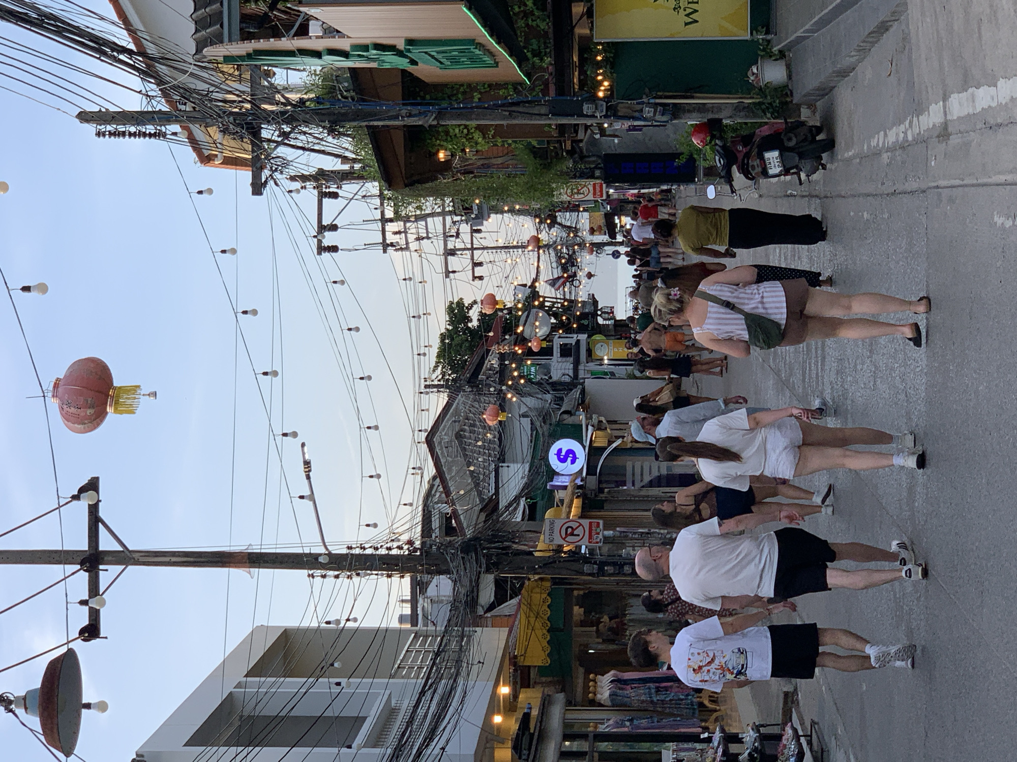 The image depicts a street scene in Chaweng, Ko Samui District, Thailand, characterized by a bustling atmosphere with numerous people walking along a narrow road lined with shops and businesses. Overhead, a dense network of electrical wires crisscrosses the sky, adorned with numerous small light bulbs and a few larger lanterns. Buildings on both sides of the street showcase a variety of architectural styles, with some featuring open storefronts displaying clothing and other merchandise. A prominent sign with a dollar sign symbol in a circle is visible on the right side of the street. A red helmet is resting on a parked scooter. The lighting suggests it is either early morning or late afternoon, with a soft, diffused light bathing the scene. The ground is paved and shows some wear and tear. The overall impression is one of a lively tourist area, with a strong emphasis on commercial activity and a vibrant street life.