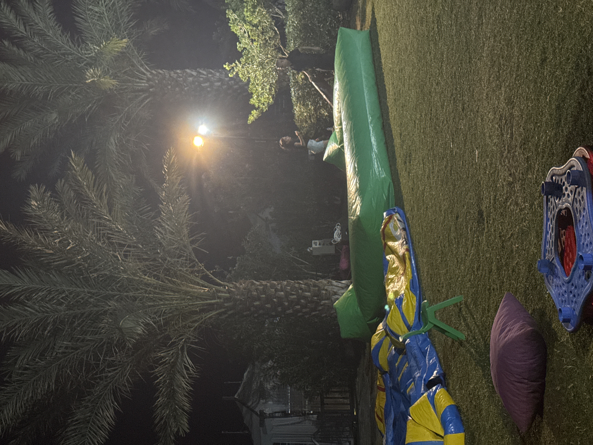 The image captures a nighttime outdoor scene, likely a backyard or park in Aksu, Turkey, featuring inflatable play structures and palm trees. In the foreground, a deflated blue and yellow inflatable structure is visible on the grass, along with a purple cushion and a partially visible blue and red play apparatus. A long, green inflatable structure, possibly a bouncy castle or obstacle course, dominates the mid-ground. Two individuals are present in the background, standing near a lit tree and a hedge, with one appearing to be holding a long pole. Palm trees are prominent in the background, and a bright light source illuminates the scene, casting a glow on the foliage and structures. The overall setting suggests an event or gathering, possibly a children's party, held in an outdoor space at night.