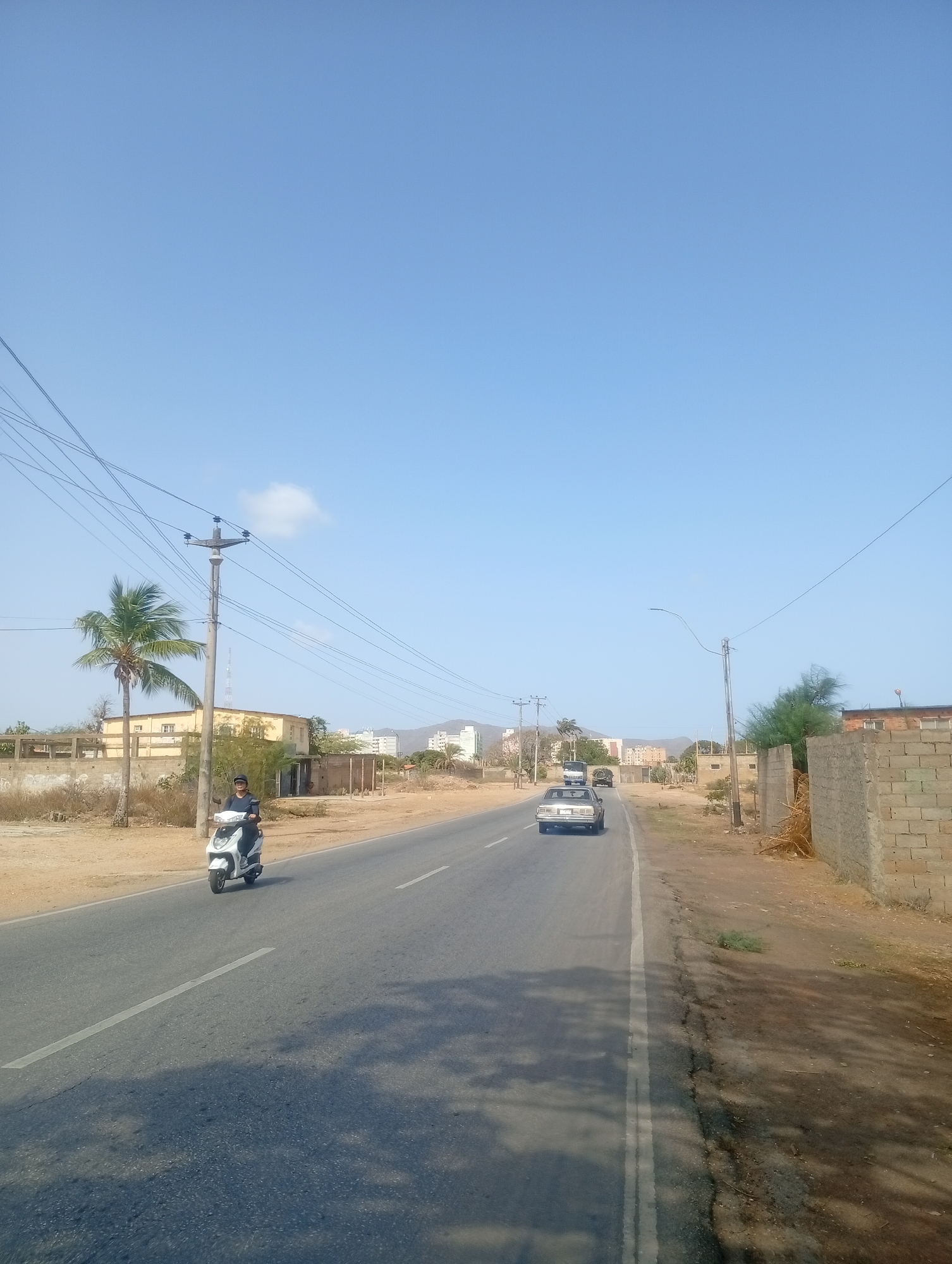 The image captures an outdoor scene on a clear, sunny day, likely in Altagracia, Venezuela. A paved two-lane road stretches through the center, receding into the distance. The road features a solid white line marking the right shoulder and a dashed white line indicating lanes.
On the left side of the road, a person is riding a white scooter, heading away from the viewer. Further down the road, a light-colored sedan is also visible, followed by a truck in the far distance. The immediate areas flanking the road consist of dry, sandy ground with sparse vegetation. To the left, a tall palm tree stands beside a utility pole with multiple power lines extending across the frame. Behind these, a taller, slender tower is faintly visible, along with various buildings and distant hills or mountains.
On the right side of the road, the dry ground continues, bordered by a low concrete block wall and some dry brush. Another utility pole with a street light arm extends over the road. In the background on this side, more buildings are scattered across the landscape. The sky above is a uniform clear blue, with a single small white cloud visible near the left utility pole. The shadows cast on the road suggest mid-day sunlight. The overall setting depicts a semi-urban or rural-urban fringe area with typical road and utility infrastructure. No visible text is discernible in the image.
