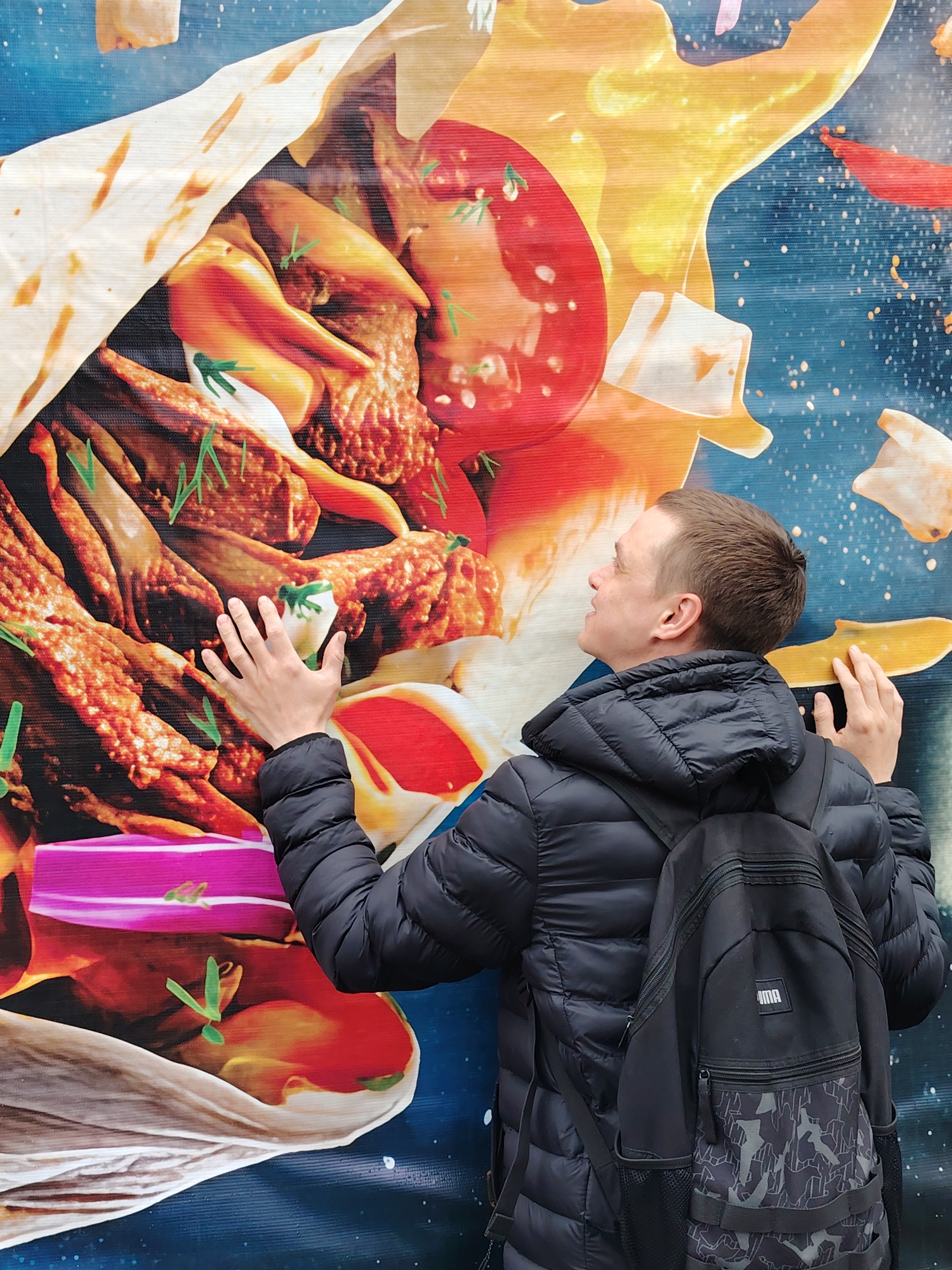 A man is photographed touching an advertisement in the city of Chaikovsky, Russia. The man is facing right and wearing a black puffer jacket and black Puma backpack. He is touching a large advertisement that features a close-up image of a shawarma or wrap. The wrap contains fried meat, tomatoes, onions, and a pink ingredient. The background of the advertisement is a blue and orange design.