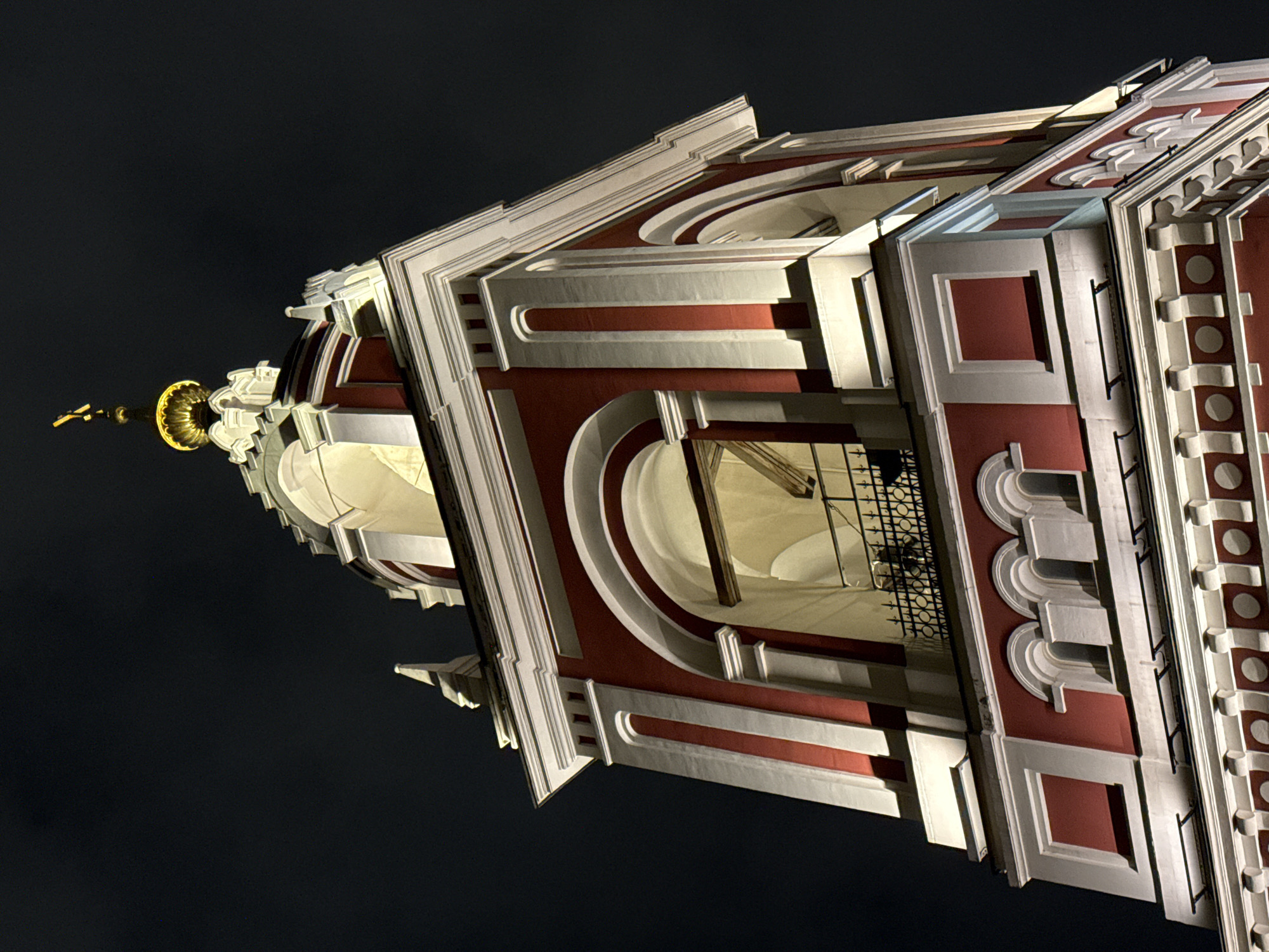 The image shows the top of the bell tower of the Church of the Life-Giving Trinity in Ostankino, Moscow, Russia, taken at night. The structure is primarily red and white with golden accents on top. The camera angle is low, looking upwards. The background is a dark sky. The bell tower features decorative elements, including rounded arches, rectangular insets, and various moldings. Metal grilles are visible inside the tower openings. The architectural style appears to be Baroque.