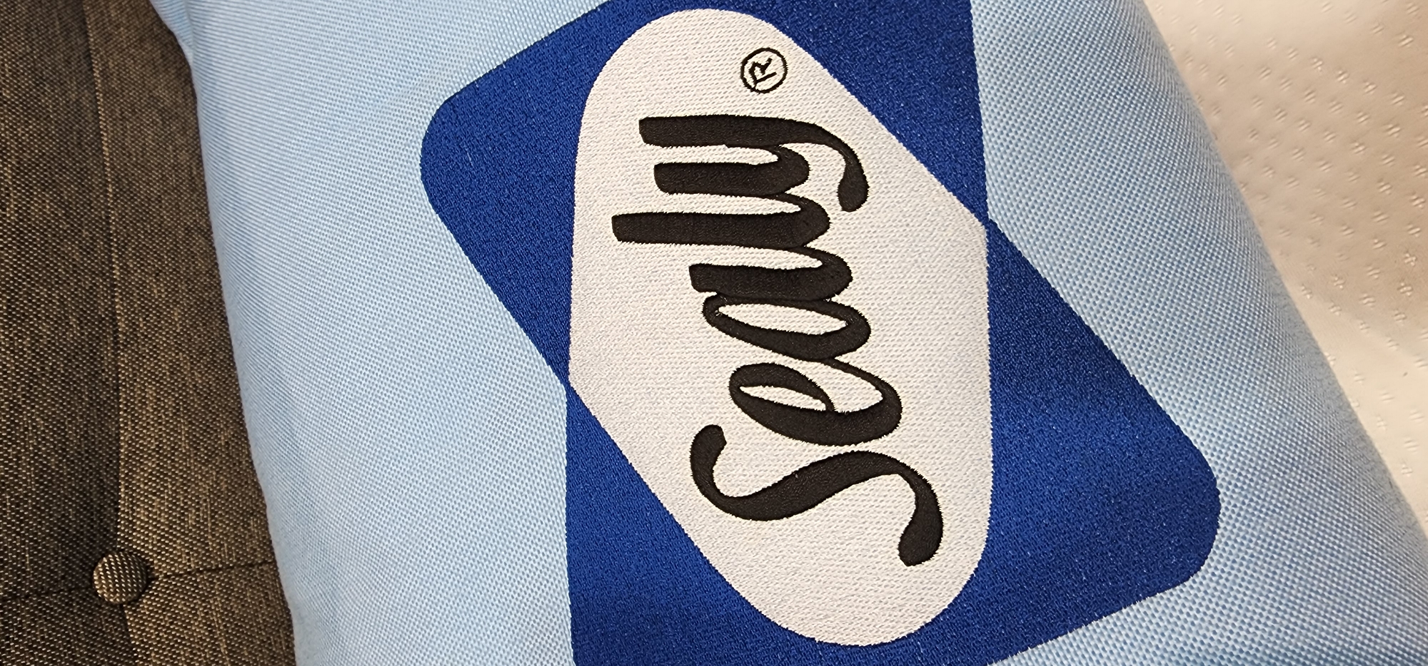 The image shows a close-up, eye-level view of a Sealy brand logo embroidered on a light blue fabric, likely a pillow or mattress cover. The logo is enclosed within a dark blue, diamond-like shape with rounded corners. The word "Sealy" is written in a cursive, black font in the center of the white embroidered oval within the blue shape. To the right of the word "Sealy" is a small, circular registered trademark symbol (®) in black. The background fabric is light blue with a visible texture, and a portion of a dark brown, textured fabric with a button is visible at the top of the image. The scene appears to be indoors, with no clear indication of its specific location within Baan Chaweng Noi, Thailand.