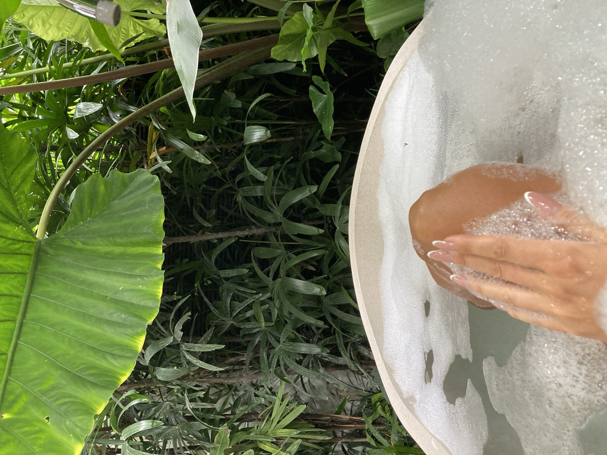 A close-up view shows a person's hands and feet submerged in a foamy bathtub, surrounded by lush green foliage. The bathtub appears to be made of a light-colored material, possibly stone or ceramic, and is filled with water and a generous amount of white bubbles. The water is a light green color. The person's hands are visible, with manicured fingernails adorned with white tips. The skin tone suggests a person of color. The backdrop is a dense array of tropical plants, with large, vibrant green leaves and thinner, elongated leaves filling the frame. Some branches are also visible among the foliage. The lighting suggests it could be daytime, possibly with dappled sunlight filtering through the leaves, creating a natural and serene atmosphere. The setting evokes a sense of relaxation and escape, possibly in an outdoor or semi-outdoor bathroom within a tropical environment. No text is visible in the image. The overall impression is one of tranquil indulgence within a natural setting. The scene takes place in North Kuta, Indonesia.