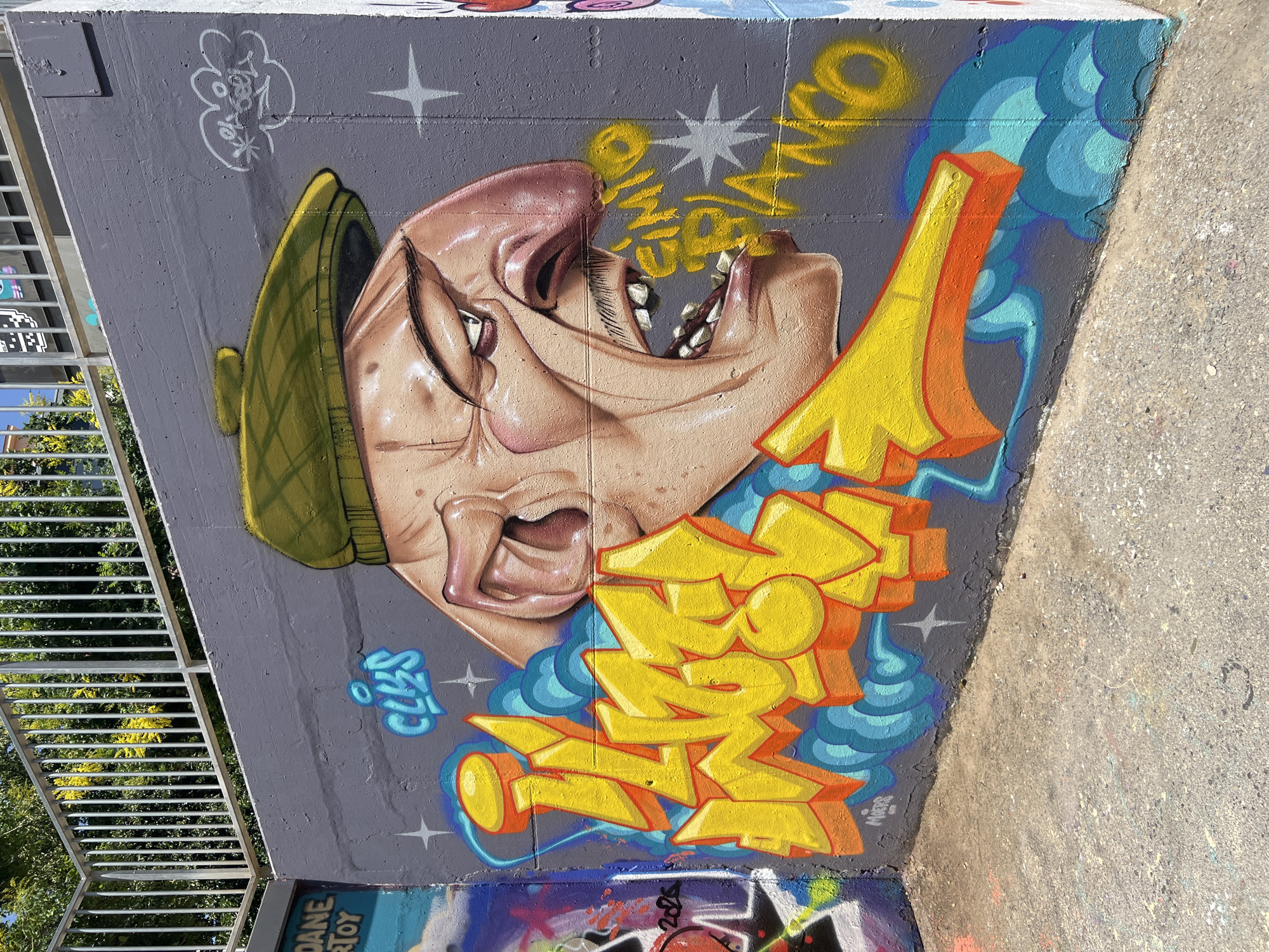 The image depicts a vibrant graffiti mural on a concrete wall in Barcelona, Spain. The mural features a cartoonish, exaggerated depiction of a man's face with a large nose and an open mouth, wearing a yellow beret. The man's expression is one of surprise or perhaps shouting. Yellow, bubble-style lettering in the foreground spells out "CLUES" in blue, and "GIRO BIANCO" in yellow is spray-painted near the man's mouth. The wall itself is gray, and the ground in front of it is rough concrete. Behind the wall, a metal railing and lush green trees are visible under a bright sky, suggesting an outdoor urban setting. The graffiti is colorful and detailed, with shading and highlights adding depth to the artwork. The corner of the wall is visible on the right side, revealing it to be a substantial concrete structure.