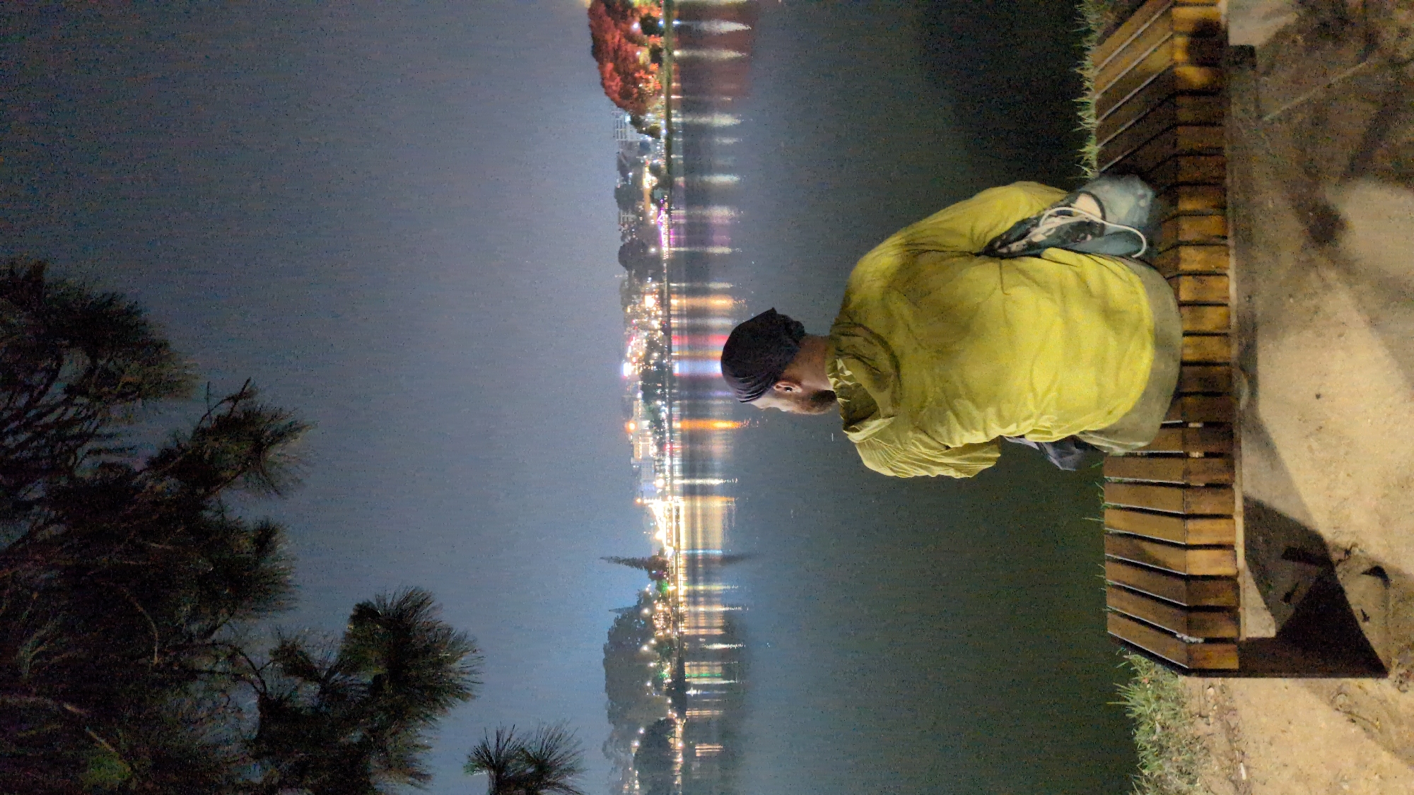 A full-body image shows a person in a yellow jacket and a black head covering sitting on a wooden bench facing a lake at night. In the background, city lights are reflected on the water, suggesting an urban area. The left side of the image features trees, contrasting with the city landscape. The person appears to be observing the scenery. The specific location is likely in or near Da Lat, Vietnam, given the reference to the city.