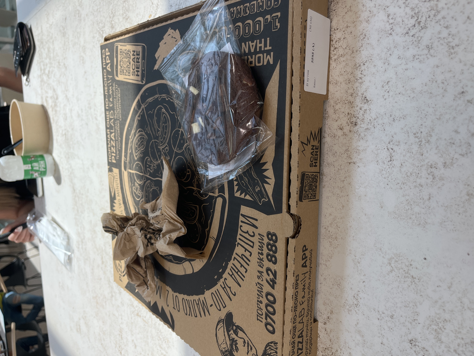 Here is a detailed description of the image:
**Scene:** The image appears to be taken on a textured, light-colored surface, possibly a table or countertop.
**Objects:**
- A pizza box from "Pizza Lab Family APP" is prominently displayed. The box features black text and graphics on a brown cardboard background. Text on the box is mostly in Bulgarian.
- On top of the pizza box, there's a wrapped brownie-like dessert in a clear plastic bag. The dessert appears dark brown with possible white chocolate chunks.
- There is a crumpled brown paper bag or napkins near the left edge of the pizza box.
- To the left of the pizza box, partially visible, are other objects.
- a black wallet, a white takeout cup and possibly other food and drink items.
- A hand is visible near a clear plastic bag which is possibly carrying utensils.
**Environment:**
- The background is partially obscured, but it appears to be an indoor setting with additional people and furniture out of focus.
**Location:** Based on the language on the box, it is likely located in Bulgaria.
**Overall Impression:** The image gives the impression of a meal or snack that has been delivered or prepared for consumption.