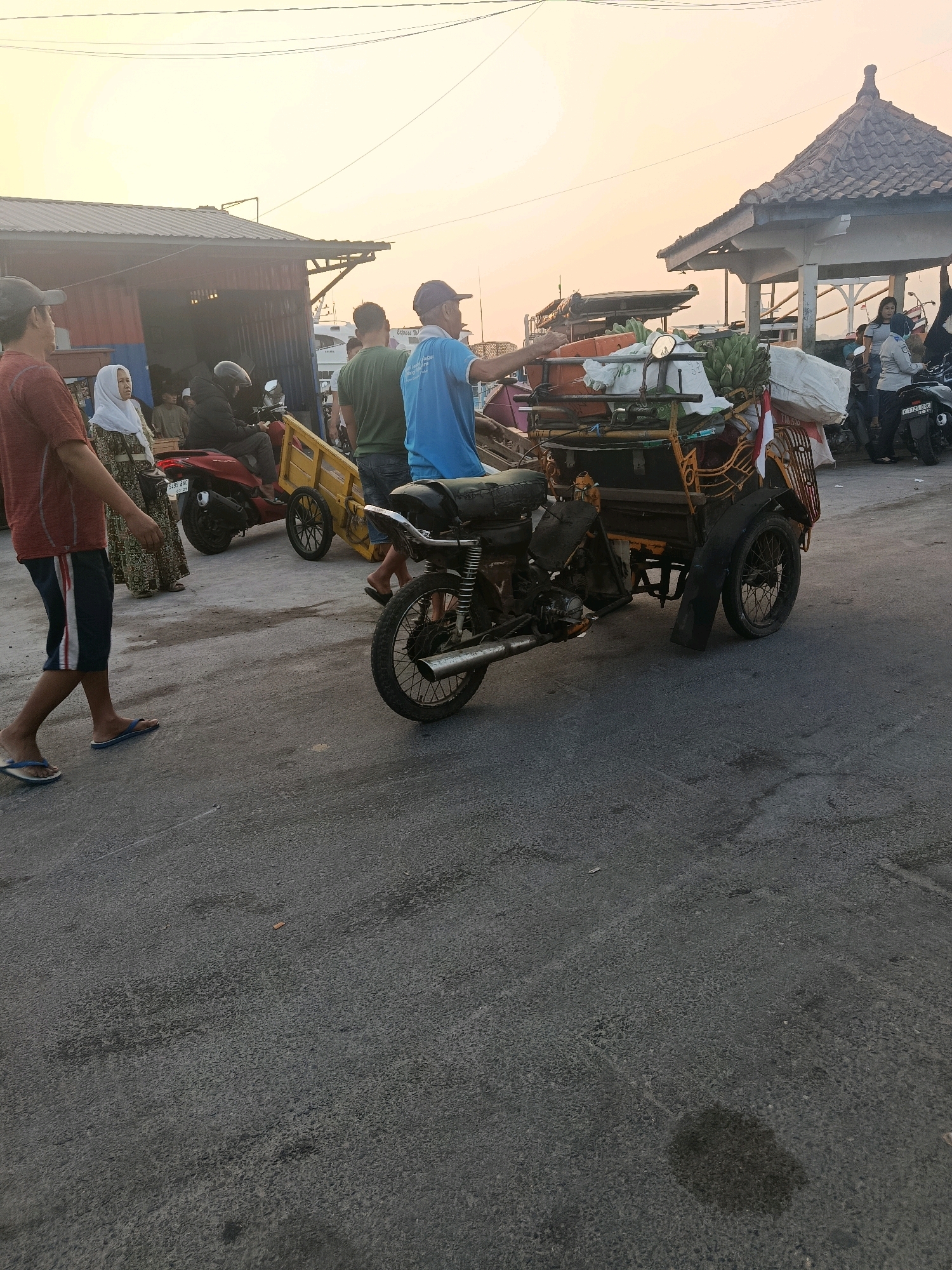 A group of people are gathered in what appears to be a market or harbor area in Jepara, Indonesia, during the late afternoon or early evening. The sky is a soft orange and yellow, indicating sunset or sunrise. In the foreground, a man in a red shirt and blue and white striped shorts walks on an asphalt road, wearing flip-flops. To his left, a woman in a patterned dress and a white hijab is standing. Behind them, a man in a black jacket rides a red scooter with a yellow cargo trailer attached.
Dominating the center of the image is a three-wheeled motorcycle with a cargo bed, laden with various items including produce like bananas and carrots, and white sacks. A person in a blue t-shirt is operating this vehicle, and another person in a green t-shirt stands nearby. A red and white Indonesian flag is attached to the back of the cargo.
In the background, a simple building with a tin roof is visible, and further back, a white gazebo-like structure with a tiled roof stands. Several other motorcycles and people are present, suggesting a busy public area. The overall scene depicts a typical day in a coastal town, likely involved in trade or transportation of goods.