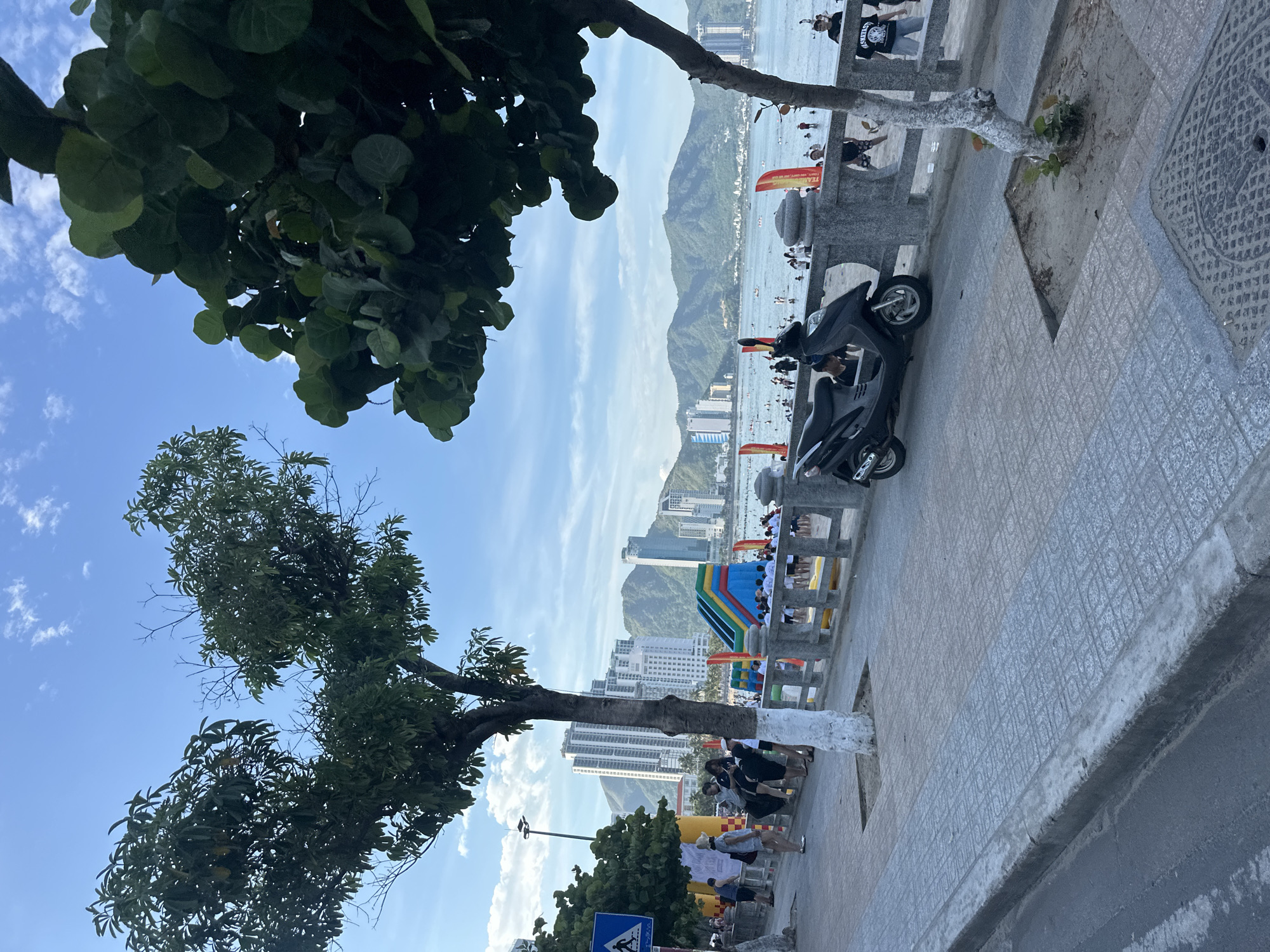 A vertical, wide-angle shot captures a bustling seaside scene in Nha Trang, Vietnam. The foreground is dominated by the lower branches and leaves of a large, leafy tree on the left, with its trunk visible, painted white at the base. To the right, a paved walkway with a patterned tile surface stretches into the distance. A dark-colored scooter is parked on the walkway.
Beyond the walkway, a stone railing separates the promenade from the beach and the ocean. The beach is populated with numerous people, some wading in the water, others relaxing. The ocean extends to a backdrop of lush, green mountains that form a dramatic silhouette against a bright blue sky dotted with scattered white clouds. In the distance, a city skyline of tall buildings rises, interspersed with palm trees.
A colorful, inflatable playground structure with slides and arches is visible on the beach, suggesting a recreational area. Red and yellow flags are placed along the promenade. The image appears to be taken during the daytime, under clear, sunny weather. The overall mood is lively and vibrant, indicative of a popular tourist destination on a pleasant day. There is a blue and white traffic sign at the bottom left with a pedestrian symbol. No other legible text is visible in the image.