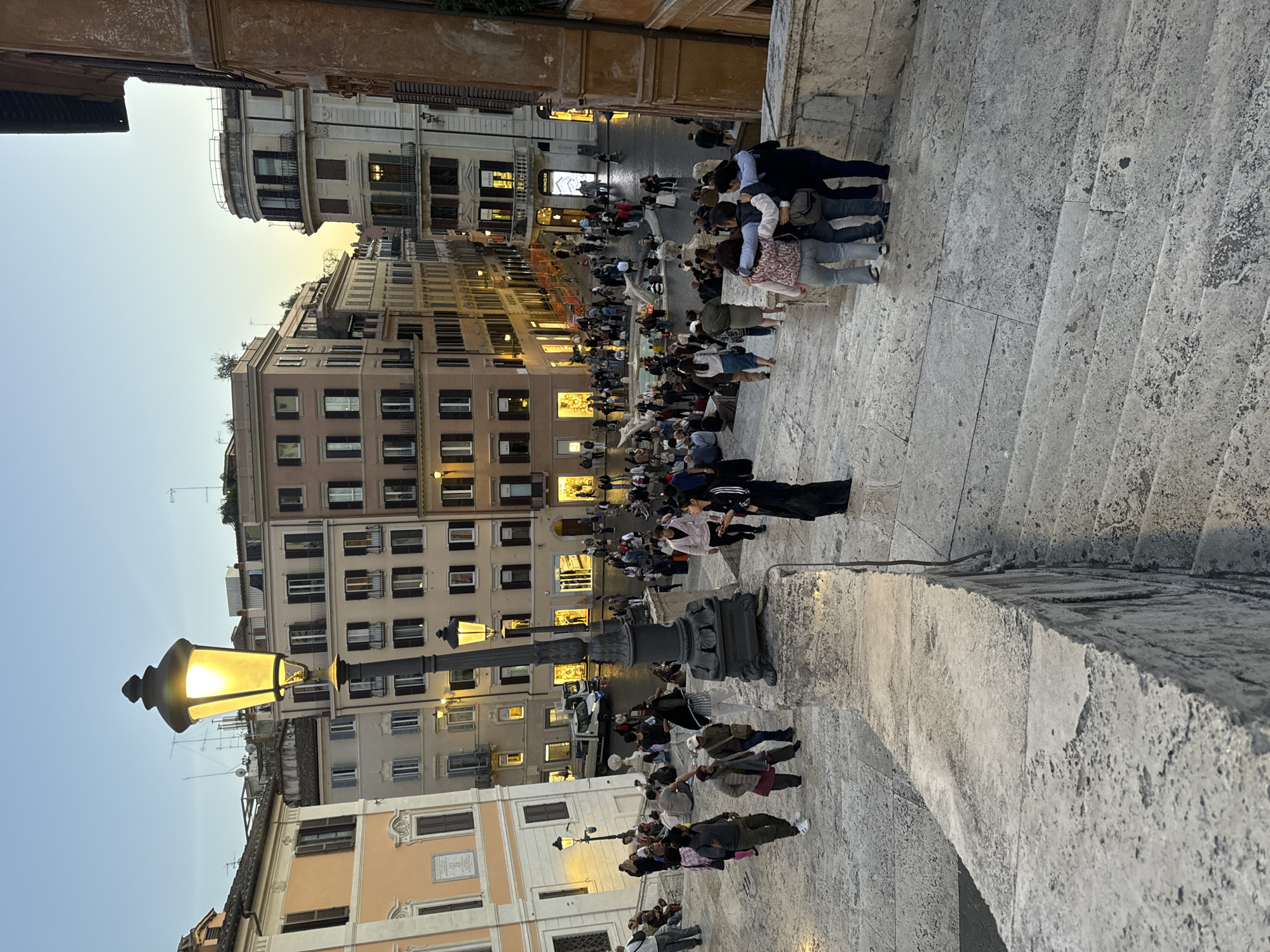 This image depicts the Spanish Steps in Rome, Italy, densely populated with people. The view is from an elevated position, looking down at the steps and the Piazza di Spagna below. Many people are seated on the steps, while others are walking through the piazza. Buildings line the background, illuminated by streetlights and the glow of dusk. A prominent street lamp stands in the foreground, its light casting shadows on the steps. People are interacting in small groups, some standing and talking, others sitting and observing the scene. The overall atmosphere appears lively and tourist-oriented.