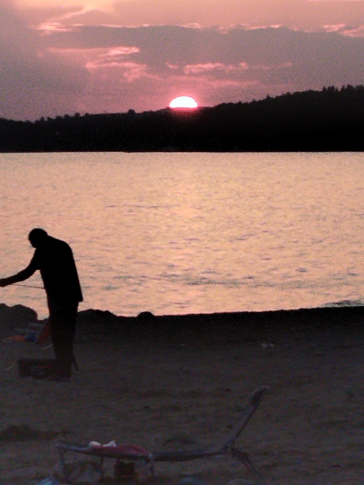An image depicts a sunset over a body of water, likely a lake or sea, with a dark, silhouetted tree line in the distance. A lone figure, also in silhouette, stands on a dark shore in the foreground, their posture suggesting they are looking out at the water. Beach chairs and other objects are scattered on the sand. The overall lighting is dim, with the sky displaying shades of pink and orange, indicative of twilight. The location is identified as Sithonia Municipal Unit, Greece.