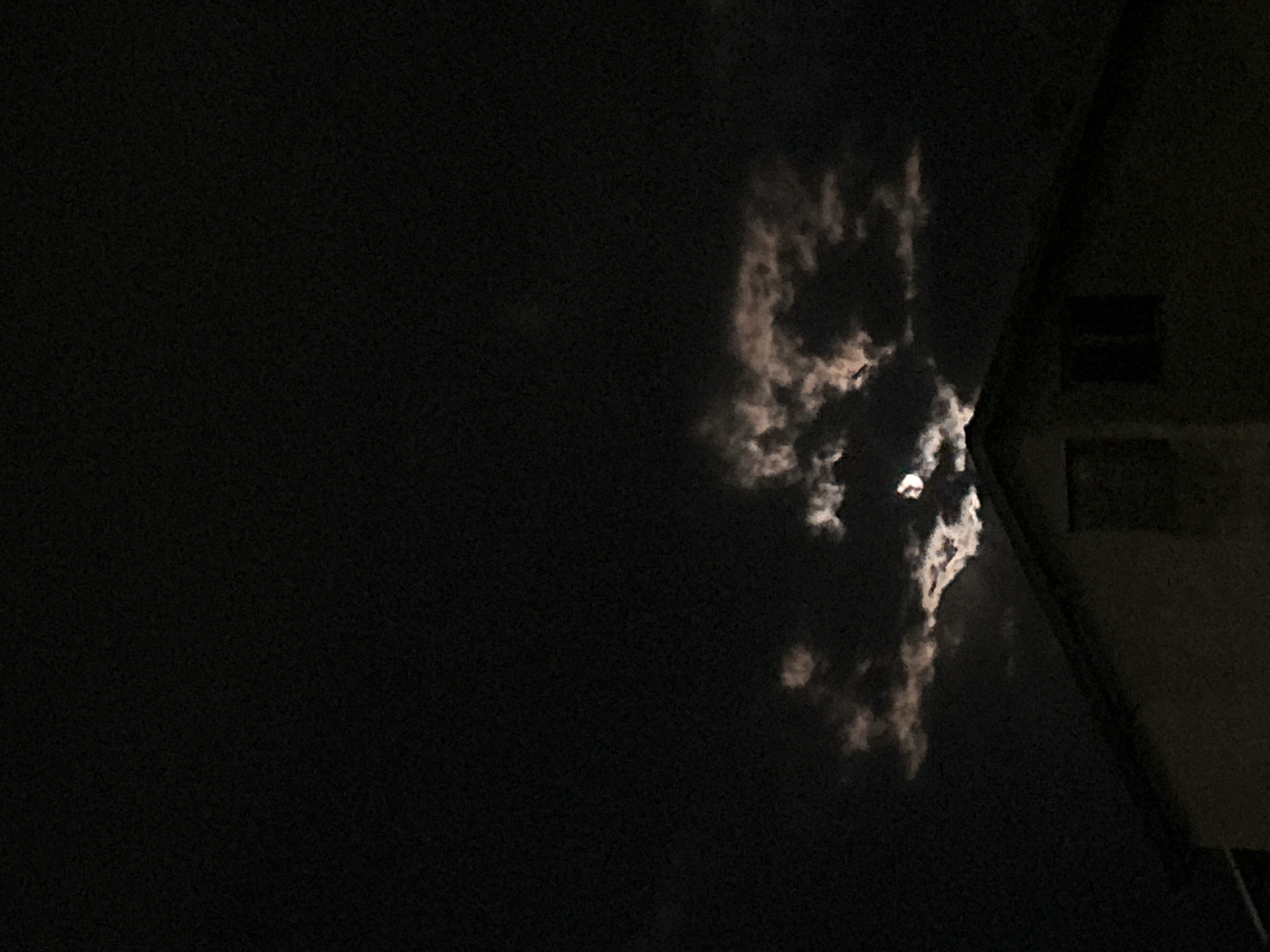 The image appears to be taken at night, with a dark sky. Bright clouds are present, with a light source behind them. The corner of a building is visible on the right side of the frame, with two dark square shapes that may be windows. There are no people or actions clearly visible in the image. The location cannot be determined to be Burgas, Bulgaria without additional information.