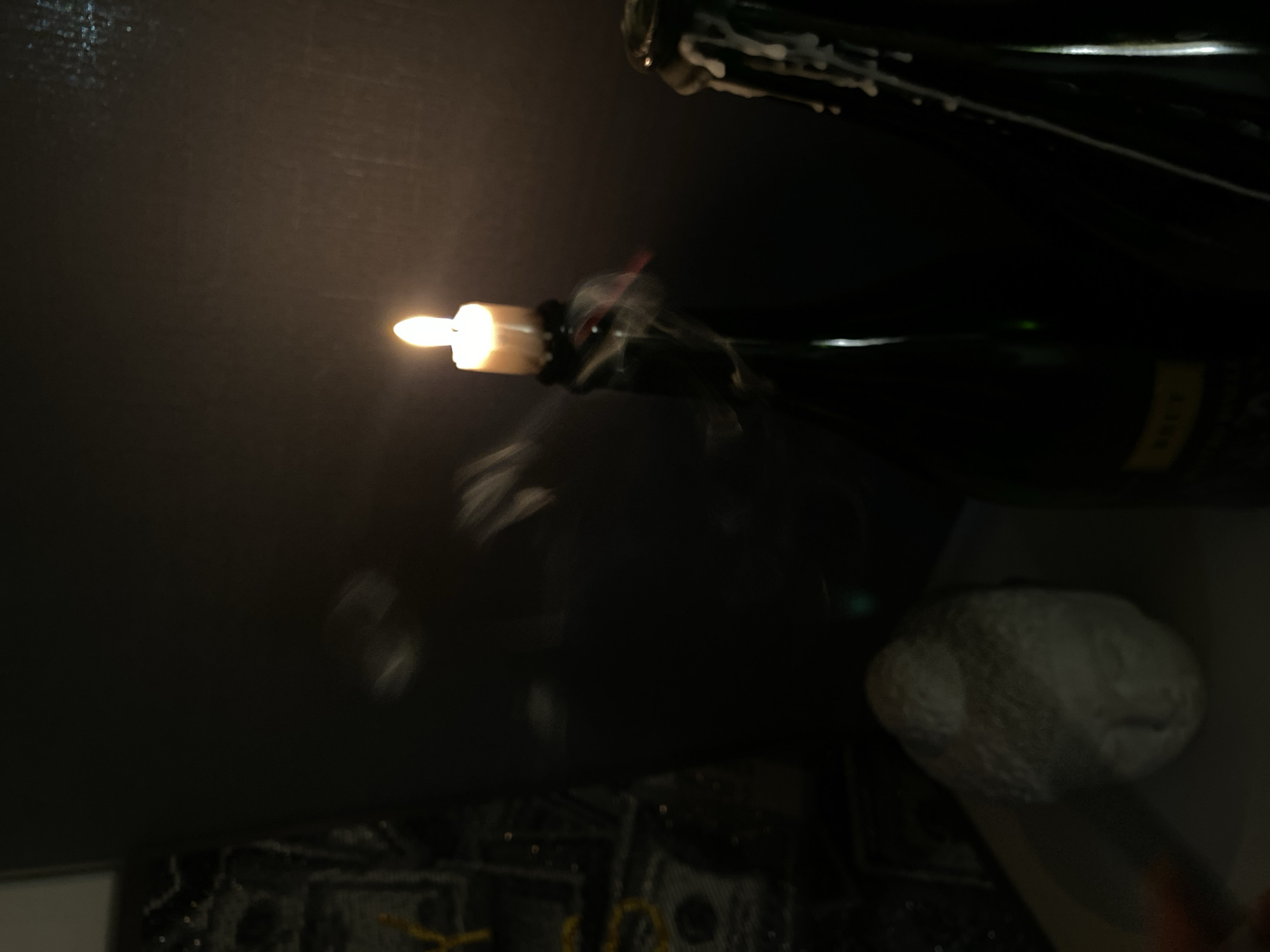 A dimly lit image shows what appears to be a partially visible champagne bottle on a dark surface, possibly glass, reflecting the light. A lit candle is inserted into the bottle neck. There's a light gray, round object near the bottle. The lower part of the scene shows some indistinct shapes and textures. There is no indication of location or people in the image.