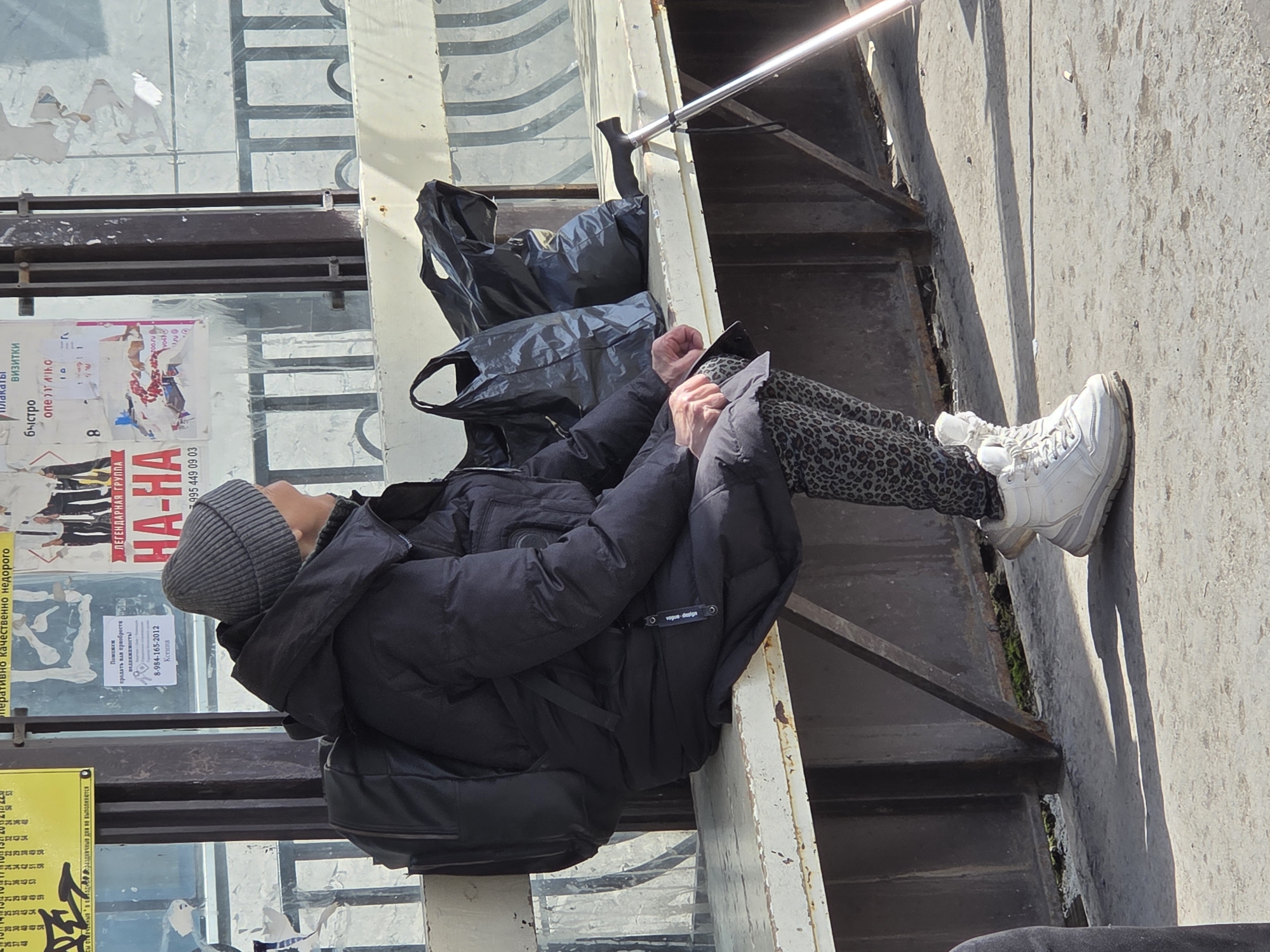 A person is sitting on the exterior metal railing of a building in what appears to be a city. The individual is wearing a dark gray beanie, a black puffy jacket, and patterned leggings, with white sneakers resting on the adjacent surface. A couple of black bags are positioned next to them on the railing. Behind the person, the side of the building is visible, covered in posters, some of which have text in Russian. The environment appears to be urban, with the building's structure showing signs of wear.