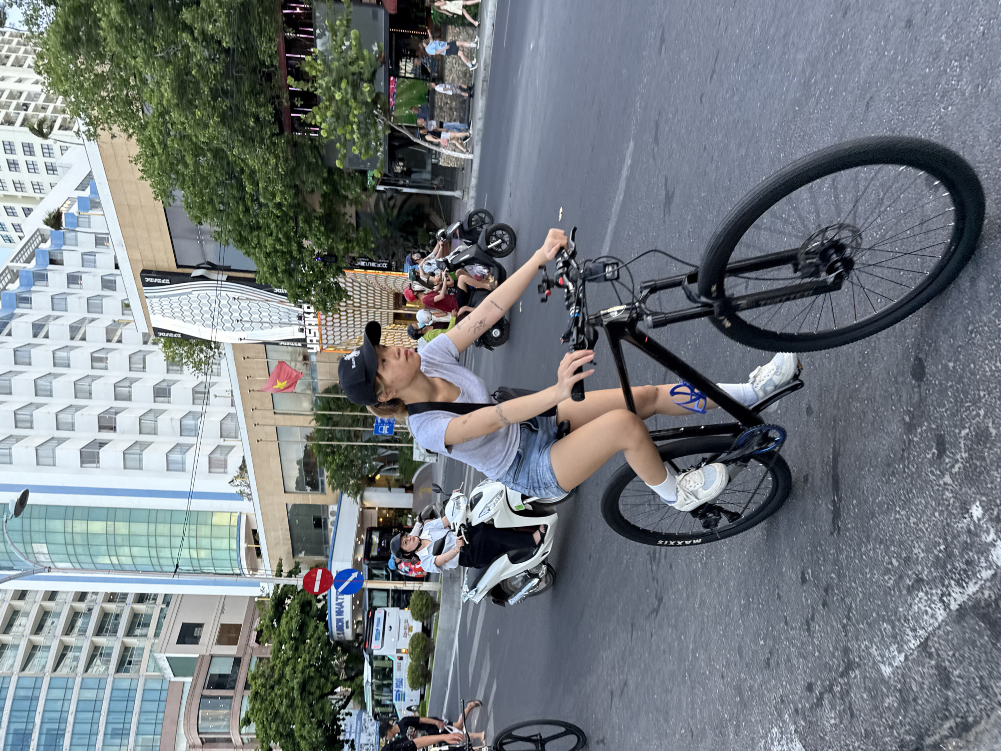 A young woman in a grey t-shirt, denim shorts, white sneakers, and a black baseball cap rides a black mountain bike on a paved road. Her right hand is on the handlebars, and her left hand is not visible. She is positioned in the foreground of the image. In the middle ground, a woman on a white scooter wearing a white collared shirt and black pants rides past. Further in the background, people walk on a sidewalk and several buildings are visible. Traffic lights and street signs are also present. The scene appears to be in a city, likely Nha Trang Ward, Vietnam, given the presence of Asian script on a sign and the style of buildings and vehicles. The lighting suggests it is daytime.