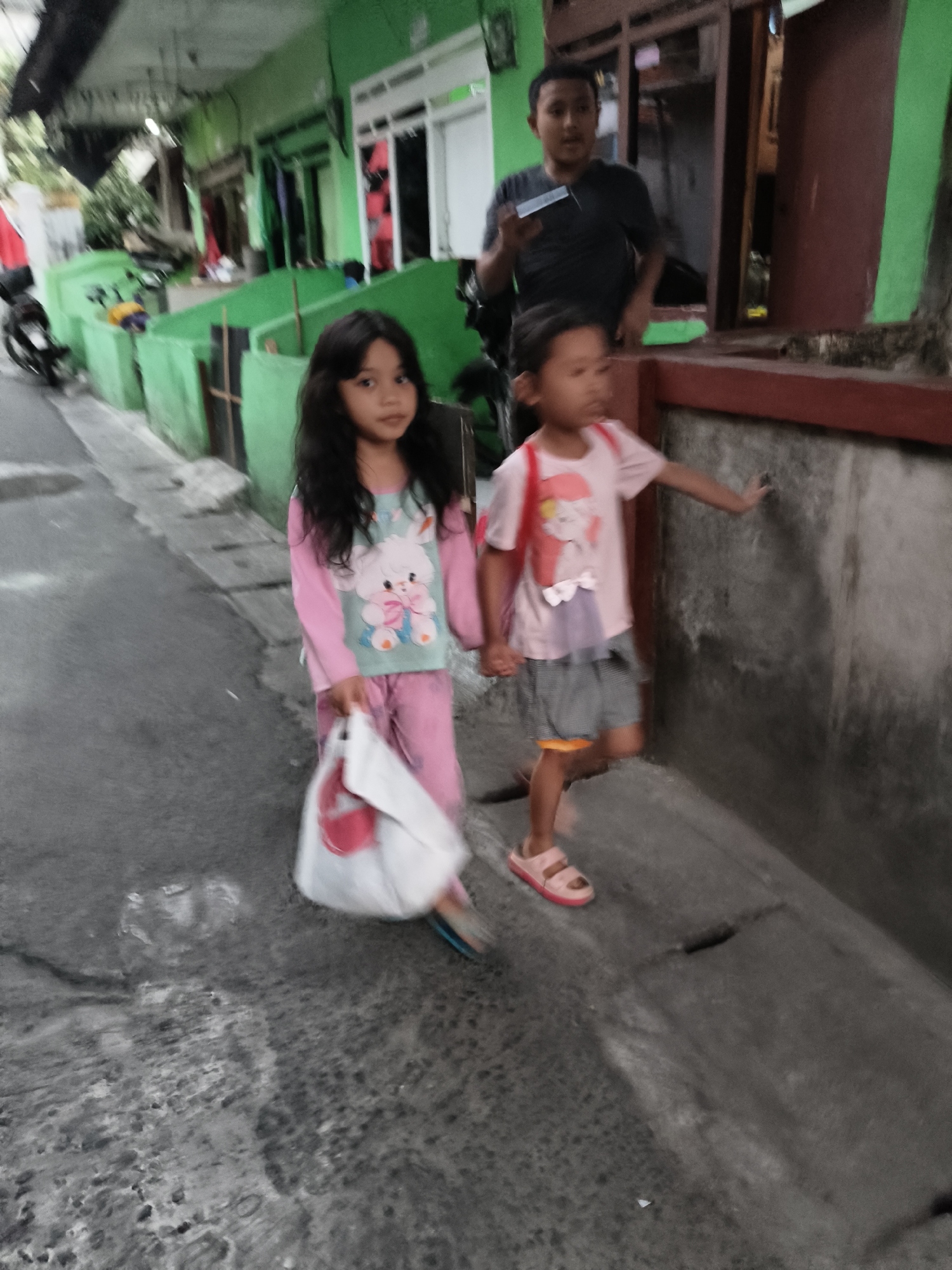 In South Tangerang, Indonesia, two young girls walk down a narrow, textured paved alley. The girl on the left, with long dark hair, wears a pink pajama set with a cartoon image of a rabbit. She carries a white plastic bag with a pink handle in her right hand. The girl on the right wears a pink t-shirt with a graphic print and gray shorts over an orange undershort. She has a small pink backpack on. Behind the girls, an adult male in a dark t-shirt stands near a building entrance, holding a dark object, possibly a phone, in his hand. The environment features green painted walls and a dark brown door on the right. There are a few motorcycles parked in the background on the left. The lighting suggests it is either early morning or late afternoon.
