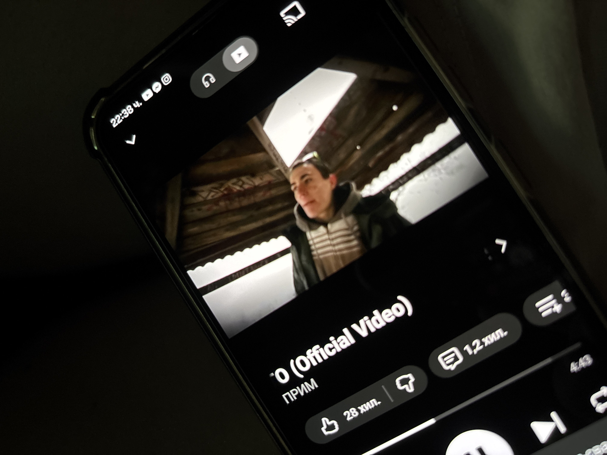A smartphone displays a YouTube video with a man visible in the frame. The video title is "O (Official Video)". Below the title is "ПРИМ". The video has 28,000 likes and 1,200,000 comments. The time displayed on the phone is 22:38. The video has 4:43 duration. The time and date imply that the location is within the city of Koprivlen, Bulgaria.