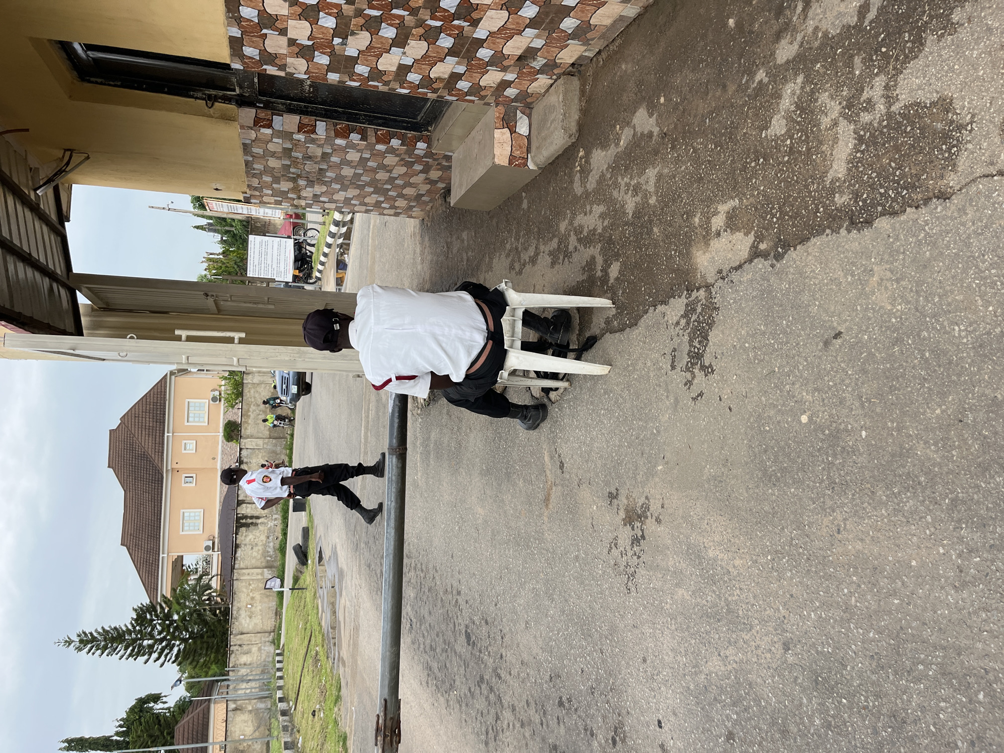 A street view in Abuja, Nigeria, presents two individuals in similar uniforms: white shirts with red accents and dark trousers. One man is seated on a white plastic chair near a building with tiled walls, while the other is walking further down the street. A pole extends into the foreground, and a building with a brown roof is visible in the background, along with signage and some vegetation. The surface appears to be a paved road.
