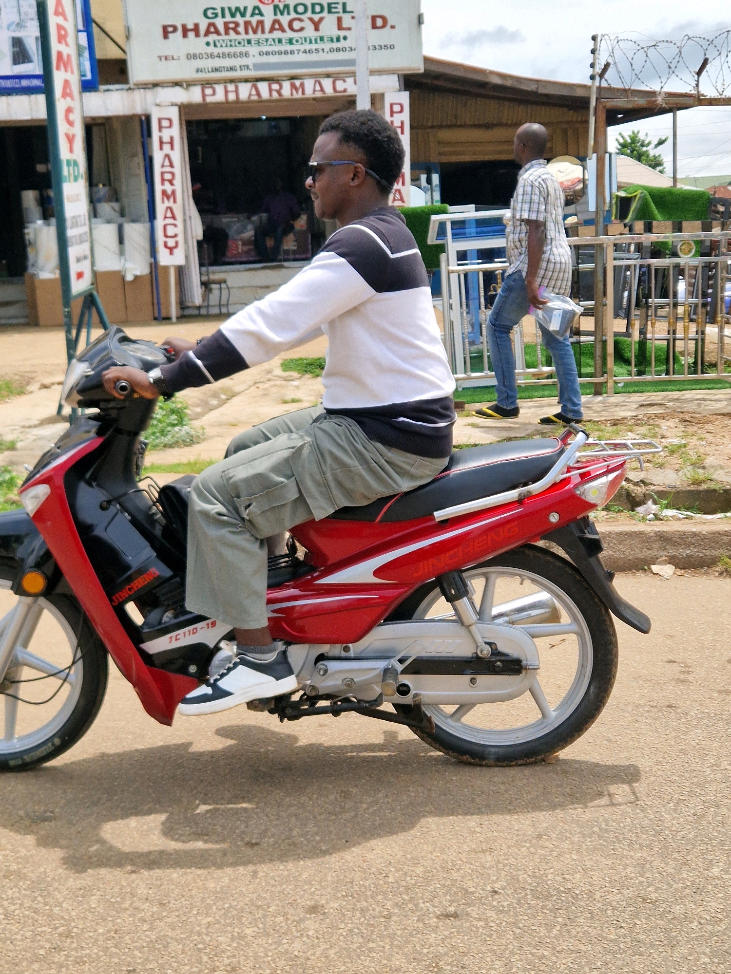 In Jos, Nigeria, a man is riding a red and black motorcycle. He is wearing a white long-sleeved shirt with black stripes on the sleeves and collar, and khaki cargo pants. He has on sunglasses and white and black sneakers. Behind him, another man in a plaid shirt and blue jeans is walking, carrying a plastic bag. The scene takes place on a street with buildings that include signage for "GIWA MODEL PHARMACY" and "PHARMACY LTD." There are fences and some greenery visible in the background, suggesting an outdoor urban environment. The motorcycle has "JINCHENG" and "JC110-19" printed on its side.