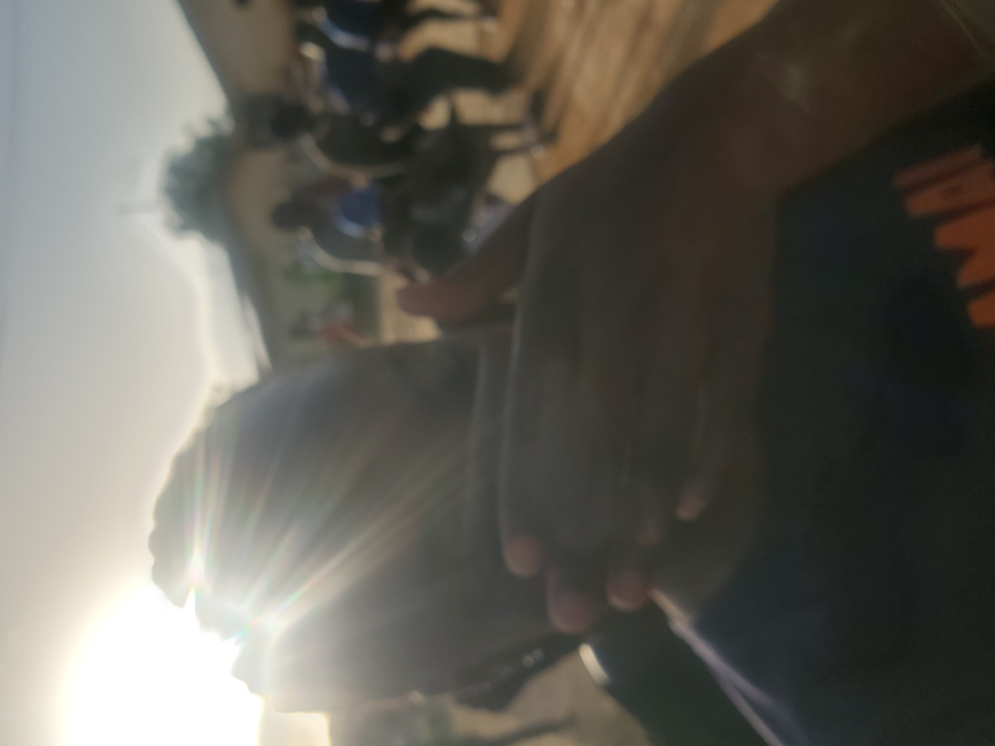 A low-angle, slightly blurred photograph shows a person in the foreground, facing away from the camera and to the left. Their head is partially obscured by bright sunlight, which creates prominent lens flare effects radiating outwards. The person appears to be wearing a dark shirt with orange lettering that is partially visible. Their hands are clasped in front of them, with one hand resting on their leg.
In the background, a group of people are visible, some of whom appear to be on motorcycles. The scene suggests an outdoor setting, possibly a street or a gathering. The bright sunlight indicates it is daytime, likely late afternoon or early morning due to the angle of the sun and the warm tones. The overall atmosphere seems casual and relaxed.
The lettering on the shirt is indistinct, but the visible orange shapes suggest it might be a logo or brand name. The context of Rafin Par, Nigeria, suggests a Nigerian setting. There are no other specific location cues or details about the weather beyond the presence of bright sunlight. The emotions conveyed are difficult to ascertain due to the blurriness and the person's pose, but the overall impression is one of a candid, everyday moment captured outdoors.