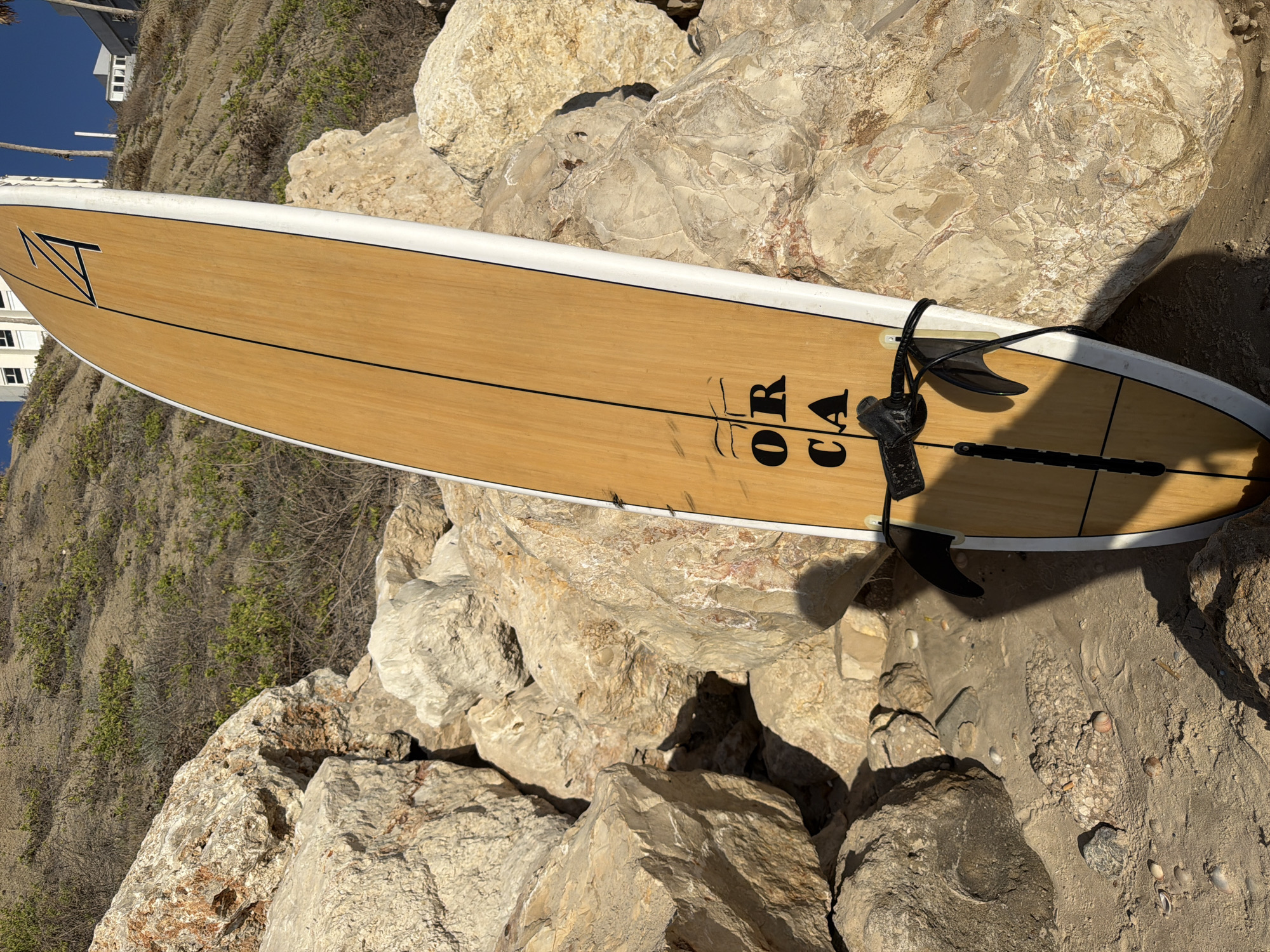 A wooden surfboard with white rails rests against a pile of large rocks on a sandy beach. The surfboard has black lettering that reads "ORCA" and a black graphic symbol resembling a stylized "Z" or "A" at the top. Two black fins are attached to the bottom of the surfboard, and a black leash is wrapped around the middle. The background shows a sandy hillside with sparse vegetation and some buildings visible in the distance. The scene appears to be a sunny day. The location is identified as Bat Yam, Israel.
