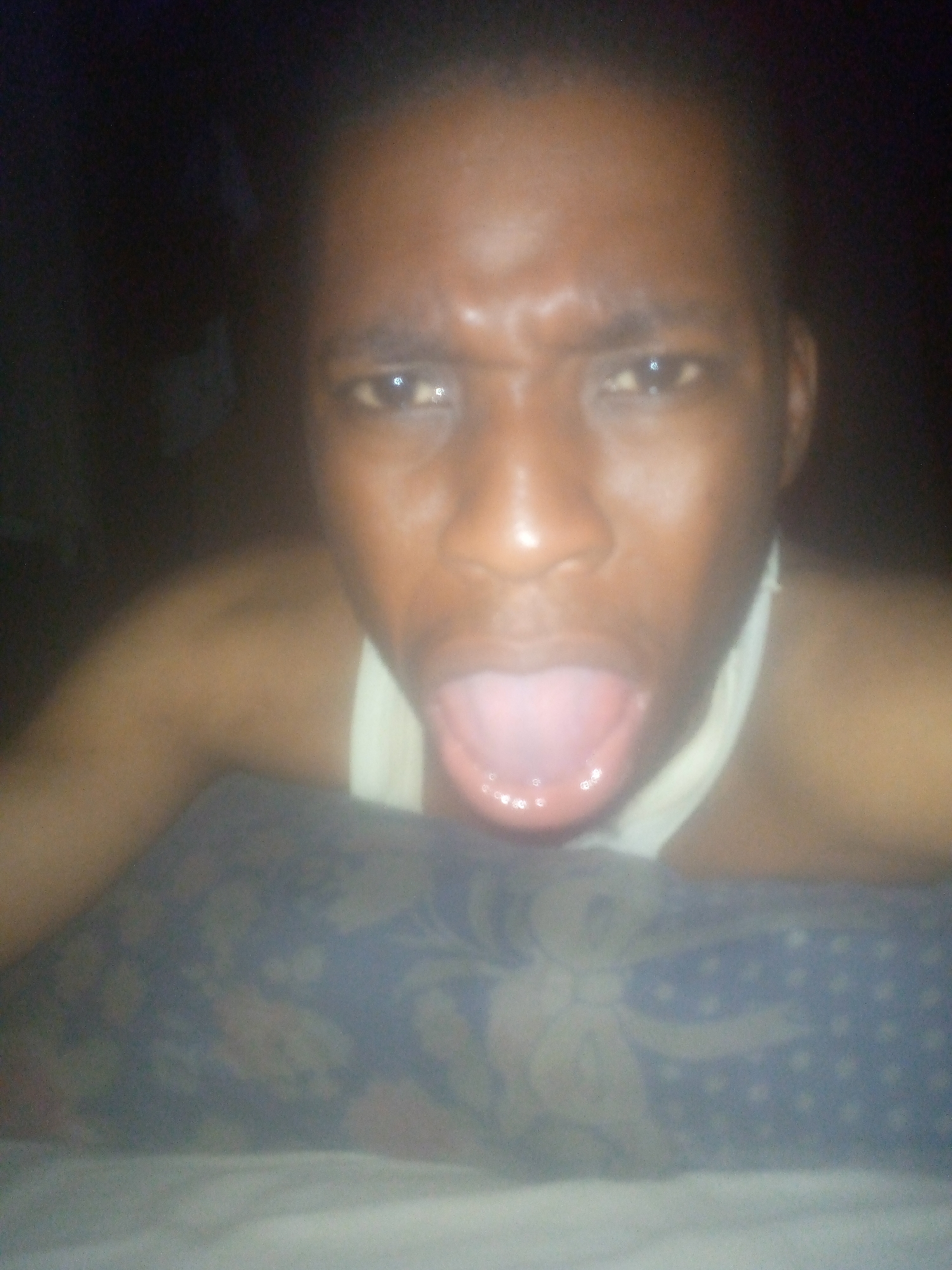 A close-up, dimly lit image captures a person, likely male, with a dark skin tone, looking directly at the camera. Their face is centered in the frame, with wide, intense eyes and furrowed eyebrows, conveying a look of surprise or strong emotion. Their mouth is open wide, tongue extended, revealing the pink interior and some visible teeth. The person appears to be wearing a white tank top or undershirt. In the immediate foreground, a pillow with a blue patterned fabric, possibly featuring floral or star-like designs, is visible. The background is dark and out of focus, suggesting low light conditions, likely evening or night. No specific location cues are discernible beyond the presence of the pillow, implying an indoor setting such as a bedroom or living room. There is no visible text in the image.
