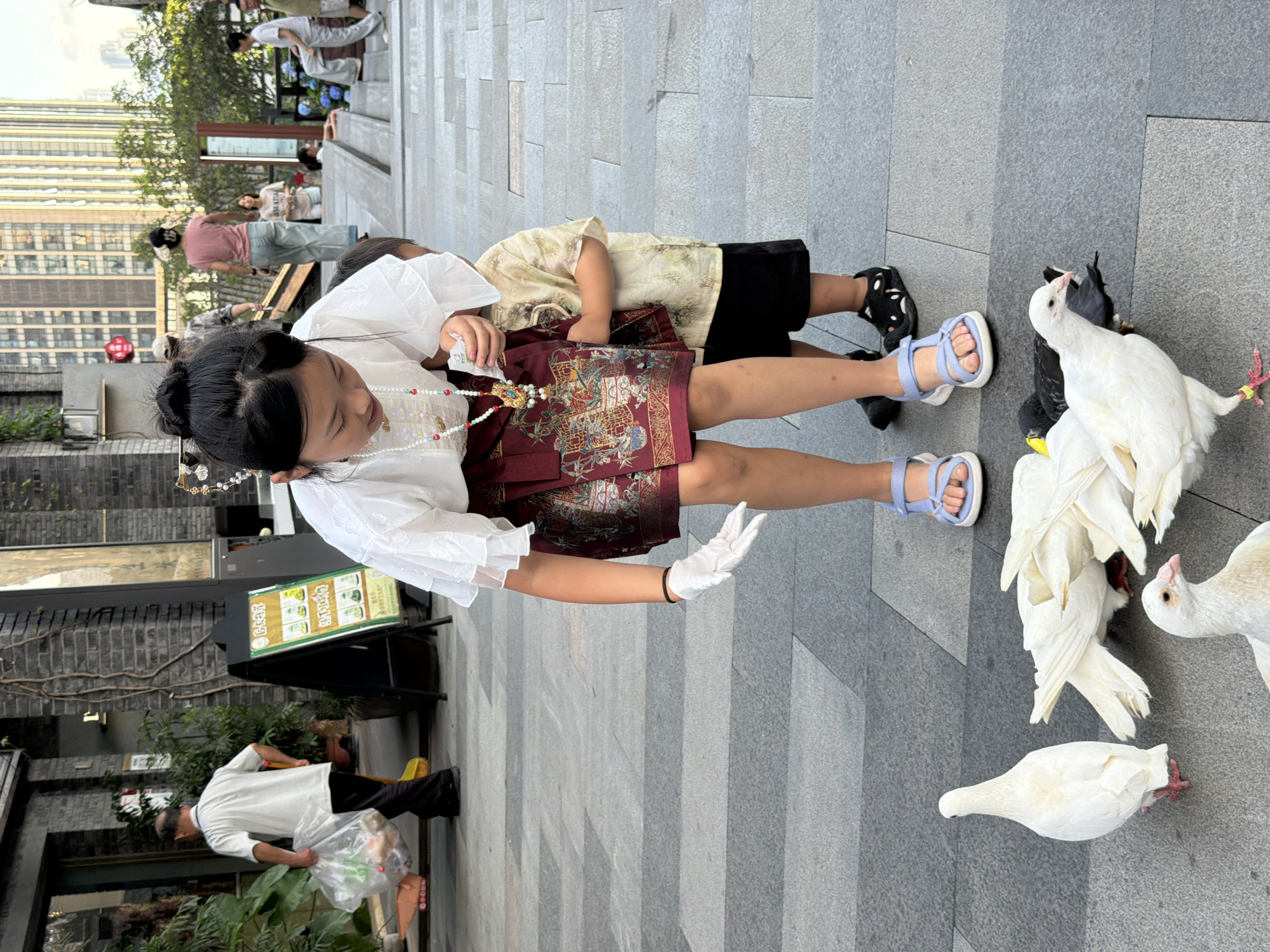 In Nan'an District, China, a young girl wearing a white top, a decorative skirt, and white gloves is crouched down, interacting with a group of pigeons on a paved plaza. She is holding something in her gloved hand, presumably food for the birds. A younger child, a boy wearing a patterned shirt and black shorts, stands behind her, partially obscured. Several white and grey pigeons are gathered around the girl's feet, pecking at the ground. In the background, a man in a white shirt and black pants walks by carrying a plastic bag, and other pedestrians are visible further back, along with buildings and greenery. The scene appears to be a public outdoor space with a modern urban setting.