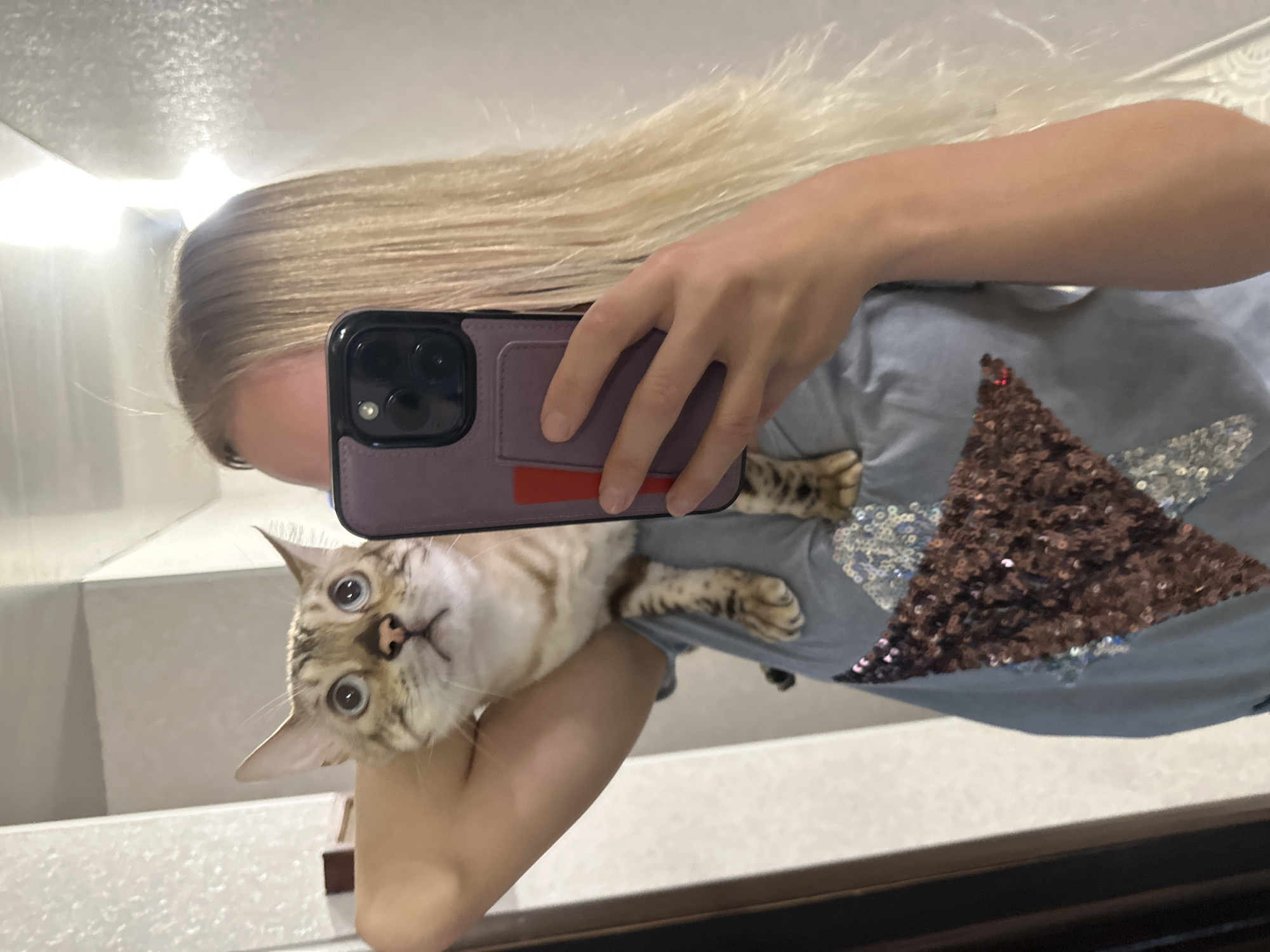 A person with blonde hair is taking a selfie in a mirror. A cat is perched on the person's shoulder. The person is wearing a blue shirt with a star made of sequins. The background appears to be an indoor setting, possibly a bathroom or hallway, with a mirror reflecting the scene. No specific location within the city of Energetik, Russia, can be identified from the image.