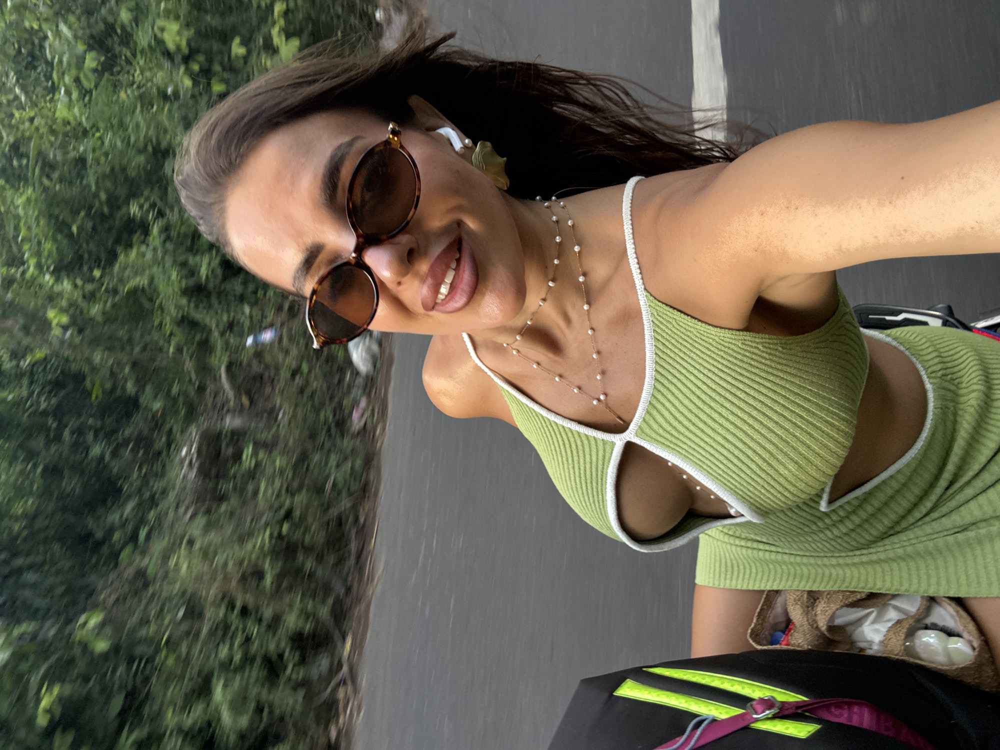 A woman is positioned at an angle, holding the camera upward. She is wearing brown sunglasses, a light green ribbed dress with a white trim, and a layered pearl necklace. She has an earpiece in her ear. Her dark hair is blowing in the wind. She is smiling. In the background, there is greenery, a gray asphalt road, and a white line marking the edge of the road. The location is likely in Kuta Selatan, Indonesia. At the bottom of the image, part of a bag with a bright yellow strip and pink detail is visible.