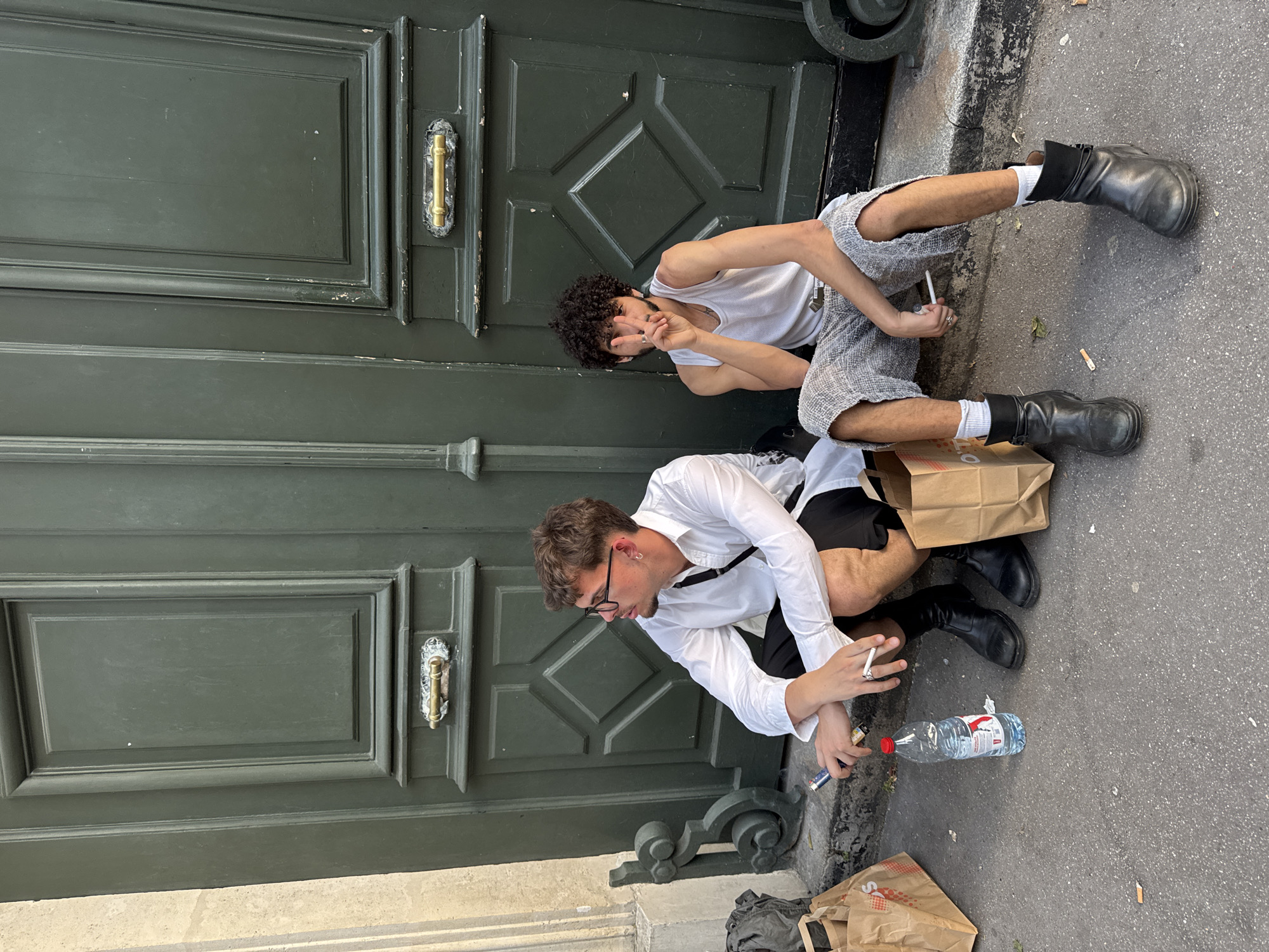 Two young men are sitting on the ground smoking cigarettes in front of a large, ornate green door. One man is wearing a white tank top and gray shorts with black boots. The other man is wearing a white long-sleeve shirt and black shorts with black boots and eyeglasses. Both men are fair-skinned and appear to be in their early to mid-twenties. A plastic water bottle and brown paper bags are on the ground near them, along with cigarette butts. The environment seems urban, possibly a residential area, in Paris, France.