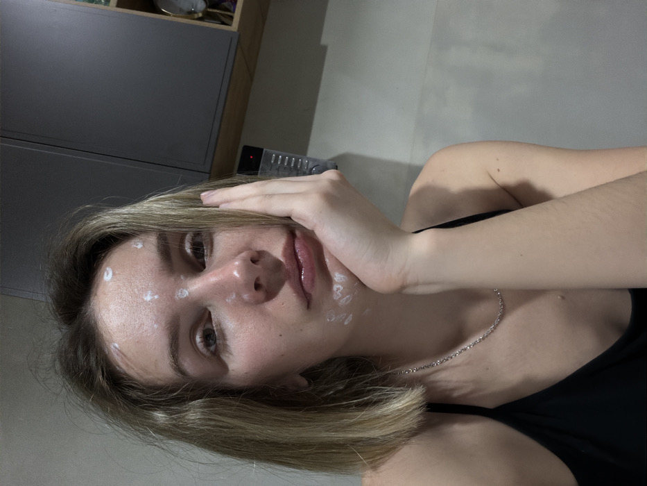 A young woman with shoulder-length blonde hair is depicted in a medium shot within an indoor setting, likely in the city of Chaikovsky, Russia. She faces the camera directly, her left hand supporting her chin. Her face, including her forehead, nose, and perioral region, has multiple distinct, circular applications of a white, pasty substance. She wears a black tank top and a thin silver chain necklace. In the background, to her right, a light-colored wall features a wall-mounted black electronic device, possibly an intercom or control panel, exhibiting a red indicator light. Above this, dark gray upper cabinets are visible, alongside a small open shelf unit containing indistinct decorative objects. The overall lighting appears soft and indirect.