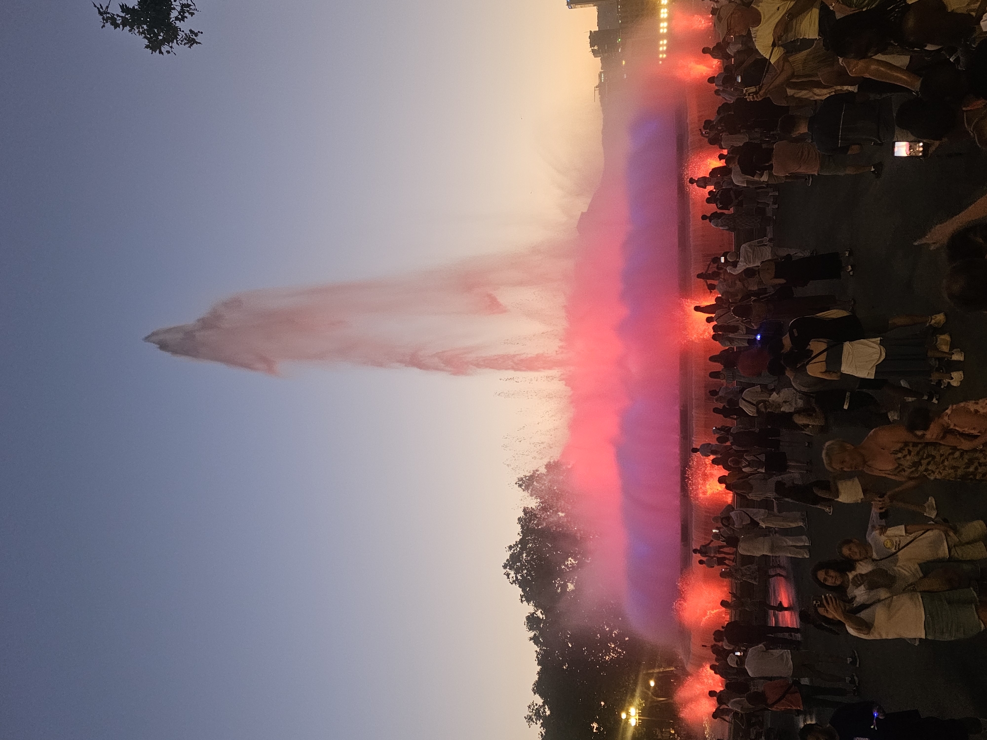 The image captures a lively scene in Barcelona, Spain, likely at dusk, given the warm, hazy sky. A large, illuminated fountain is the central focus, with water jets spraying high into the air and illuminated in vibrant shades of red and purple. The spray from the fountain creates a hazy, ethereal effect.
A crowd of people is gathered to watch the fountain show. They are standing in rows, many holding up their phones to take pictures or videos of the spectacle. The atmosphere appears excited and engaged, with individuals looking towards the fountain, some smiling. The time of day suggests a popular evening event. The city's skyline is visible in the background, with buildings illuminated by lights, hinting at an urban setting. The presence of trees suggests the fountain is located in a park or public square. The weather seems clear, allowing for an unobstructed view of the sunset colors and the fountain's display.
