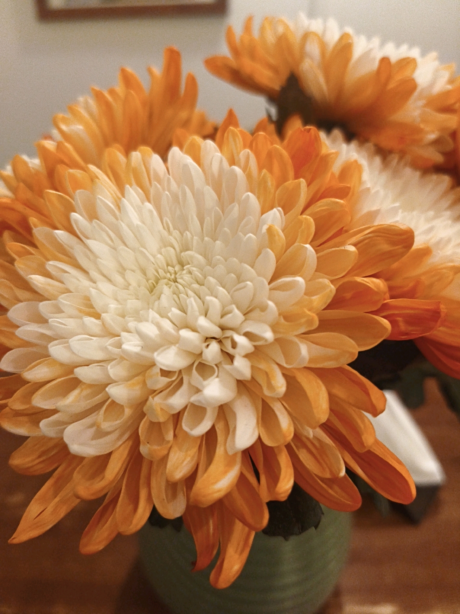 A close-up shot focuses on the intricate petals of orange and white chrysanthemums arranged in a green vase. The petals are layered and create a dense, textured bloom. The background is softly blurred, showing a warm-toned surface and a hint of a light-colored wall with a framed object. The lighting highlights the delicate details and vibrant colors of the flowers.