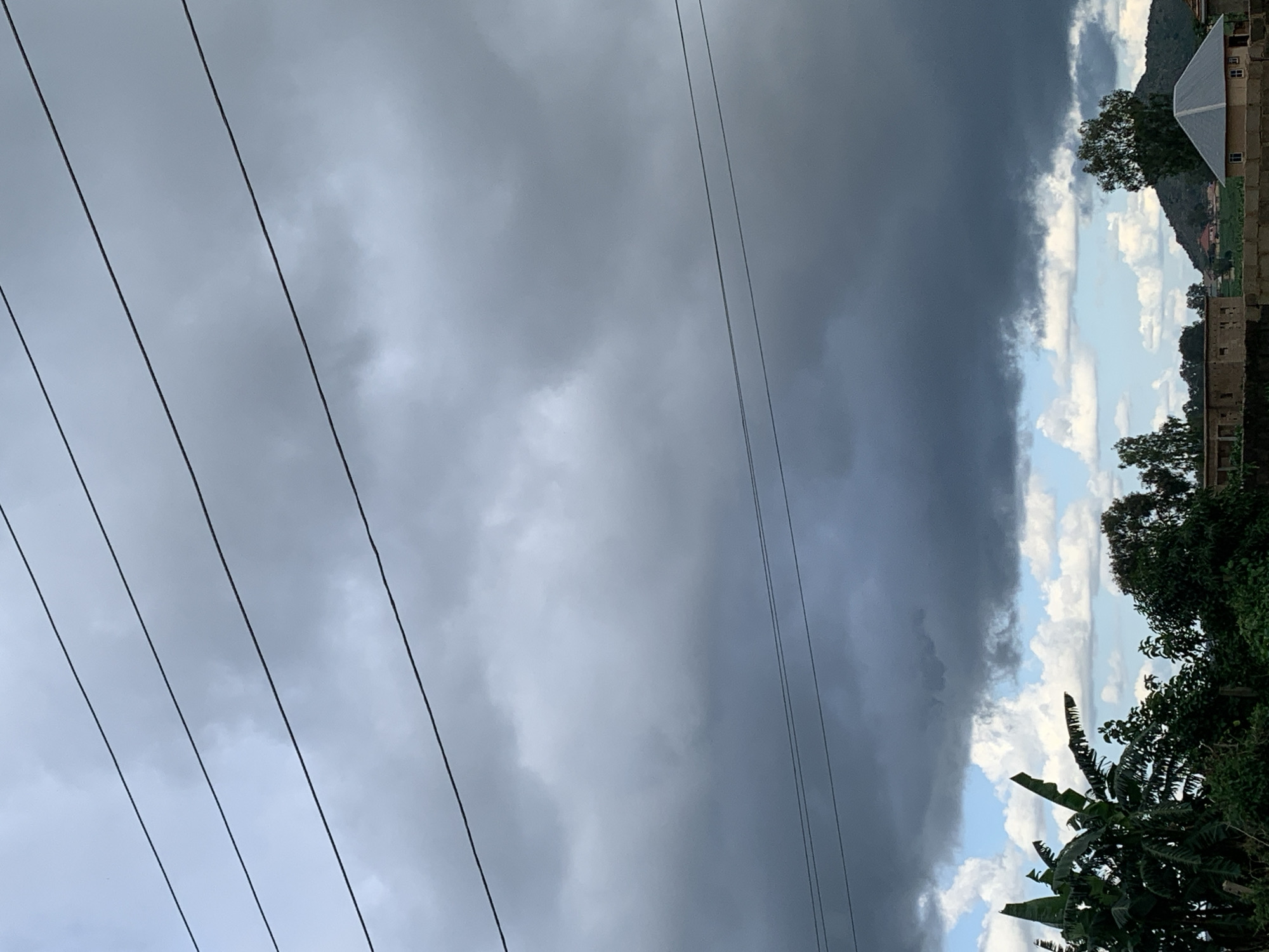 In Kuru Karama, Nigeria, the sky is dominated by dark, heavy clouds, suggesting an impending storm. Four parallel utility wires stretch across the frame, obscuring the upper portion of the cloudy sky. Below the dense cloud cover, patches of lighter, white clouds are visible against a lighter blue sky. In the foreground, lush green foliage, including banana trees, thrives, with dense vegetation filling the lower left quadrant of the image. To the right, a small, grey, roofed building stands, with a distant, dark, conical hill visible behind it. Scattered houses and trees are also present in the mid-ground and background. The overall scene captures a moment of atmospheric intensity before a potential rainfall in a semi-rural Nigerian setting.