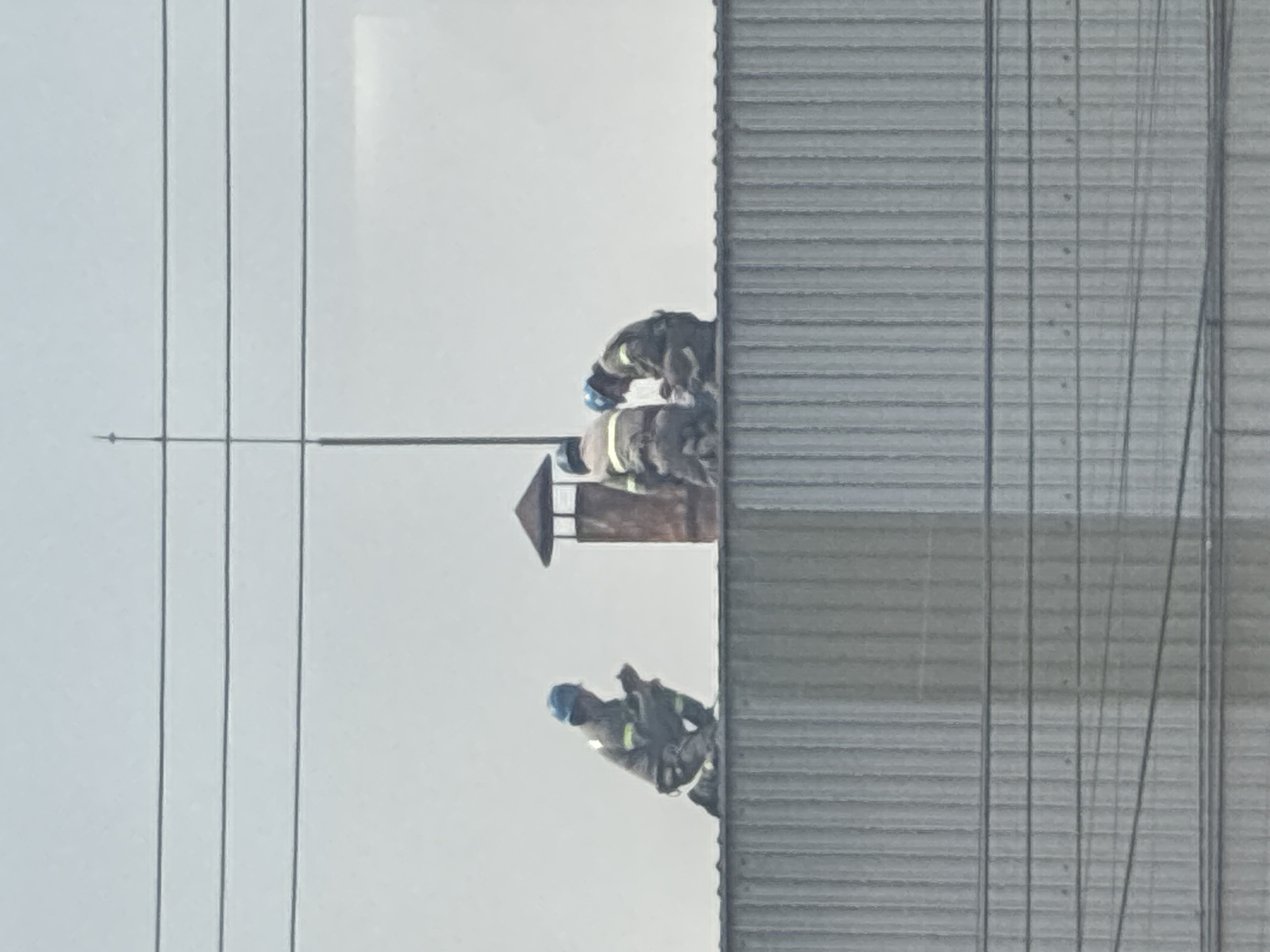 The image depicts three firefighters on the roof of a corrugated metal building. They wear blue helmets and protective gear. One firefighter is near the edge of the roof, while the other two are positioned closer to a chimney or vent. Power lines are visible in the foreground. The image was taken in Cuautitlán Izcalli, Mexico.