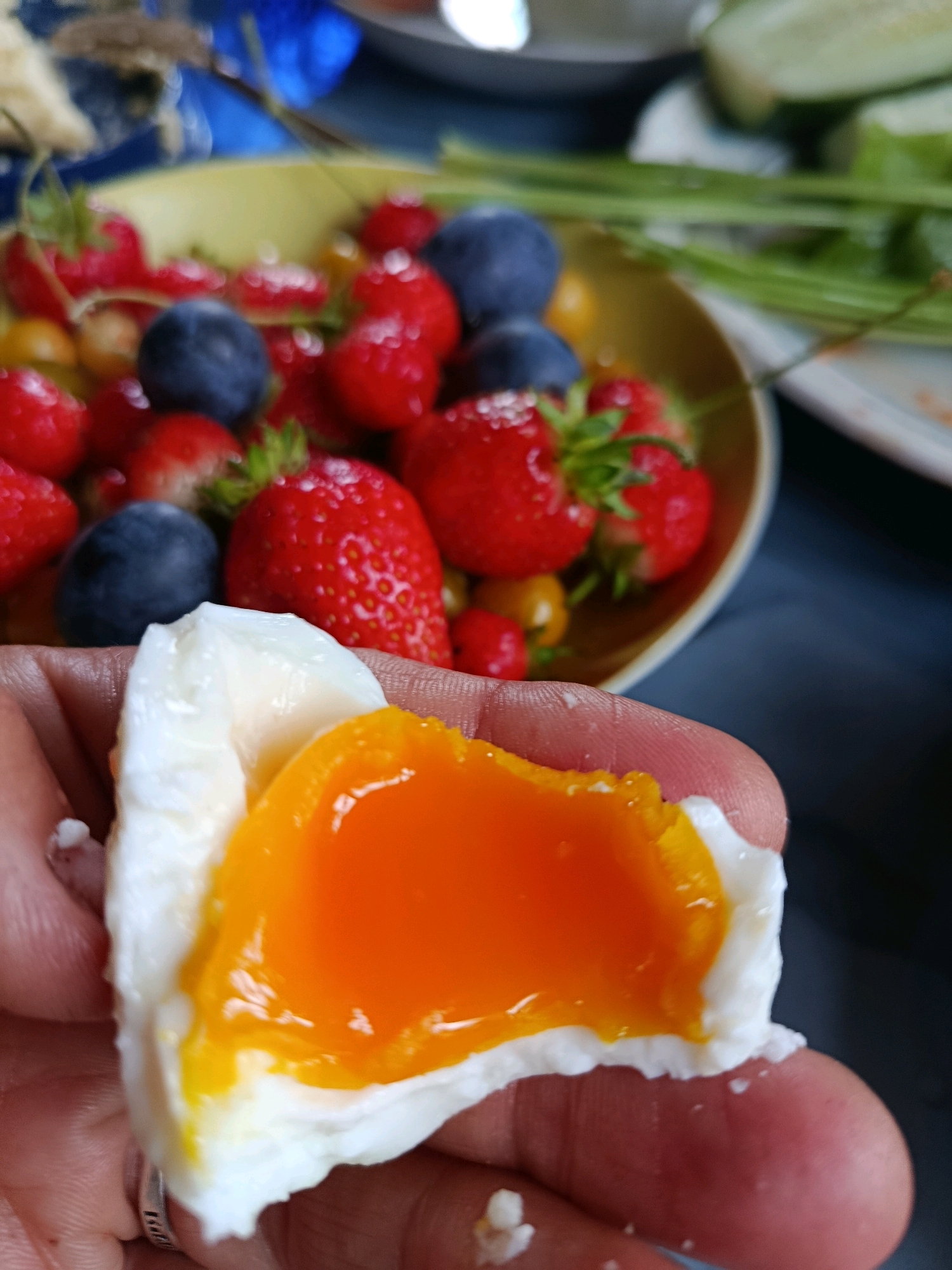 A close-up shot shows a person's hand holding a halved soft-boiled egg with a bright orange, runny yolk and white albumen. In the blurred background, a bowl of fresh strawberries and blueberries is visible, suggesting a meal or snack. The lighting is natural, and the focus is on the egg.