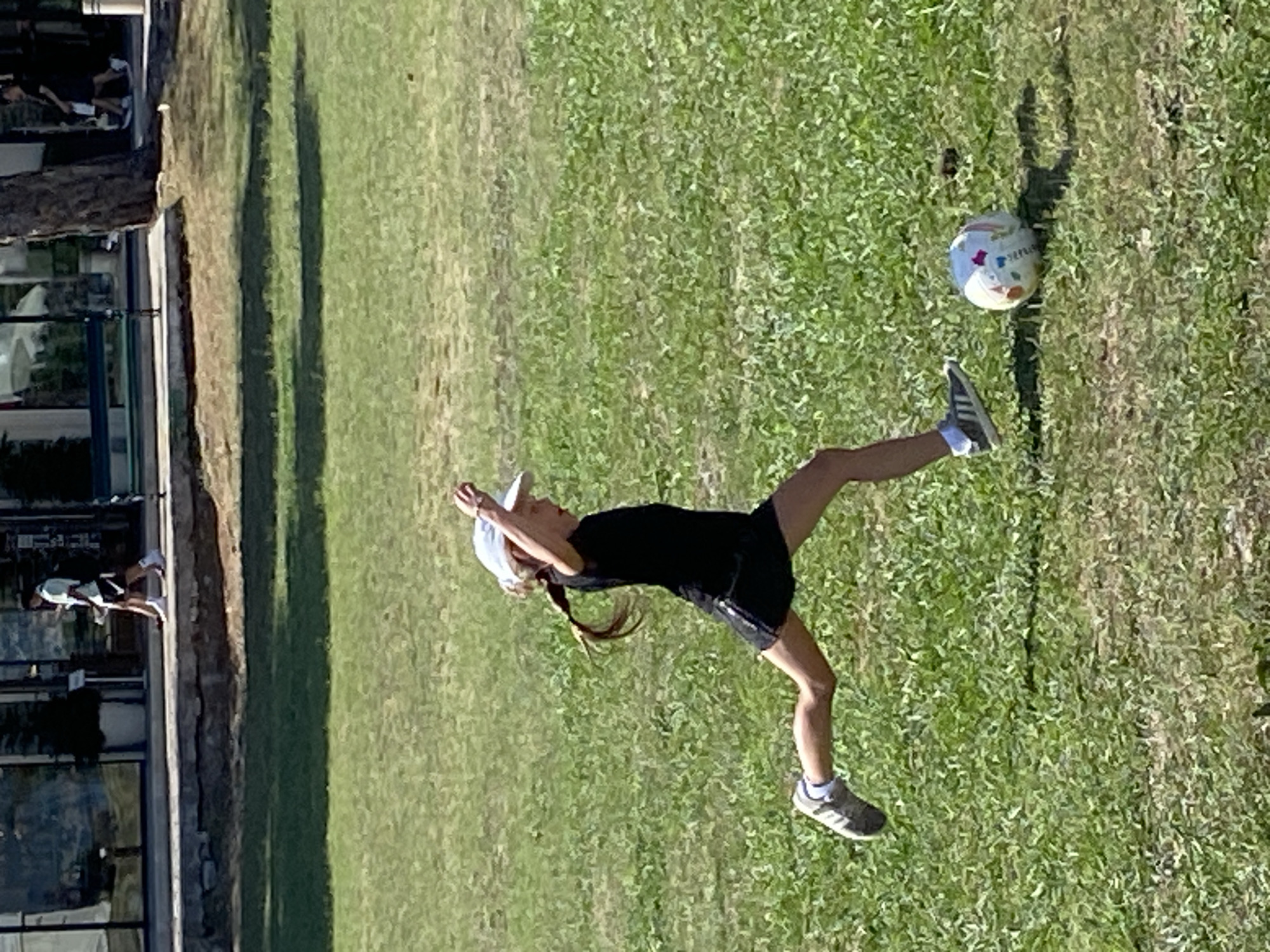 A young girl in a black t-shirt, black shorts, and a white baseball cap with her hair in a braid is kicking a colorful soccer ball on a grassy field. She is in mid-stride with her right leg extended, about to make contact with the ball. The scene appears to be a park or recreational area with a sidewalk and buildings in the background. The lighting suggests it is daytime with the sun casting shadows on the grass. The context of Budva, Montenegro is not discernible from the visual information alone.