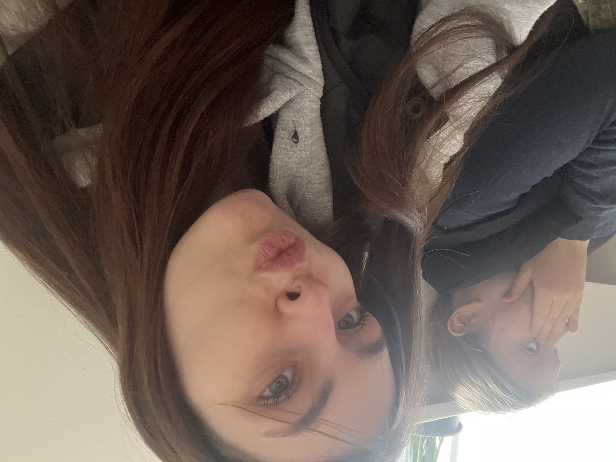 Two young women are pictured indoors. One is oriented toward the camera, making a kissing face. She has long brown hair, light skin, and blue eyes, and she is wearing a gray hooded sweatshirt over a black garment. The other young woman is seated at an angle to the camera and obscuring her face with her right hand. She has blonde hair. The background is a white wall. The location is not discernible.