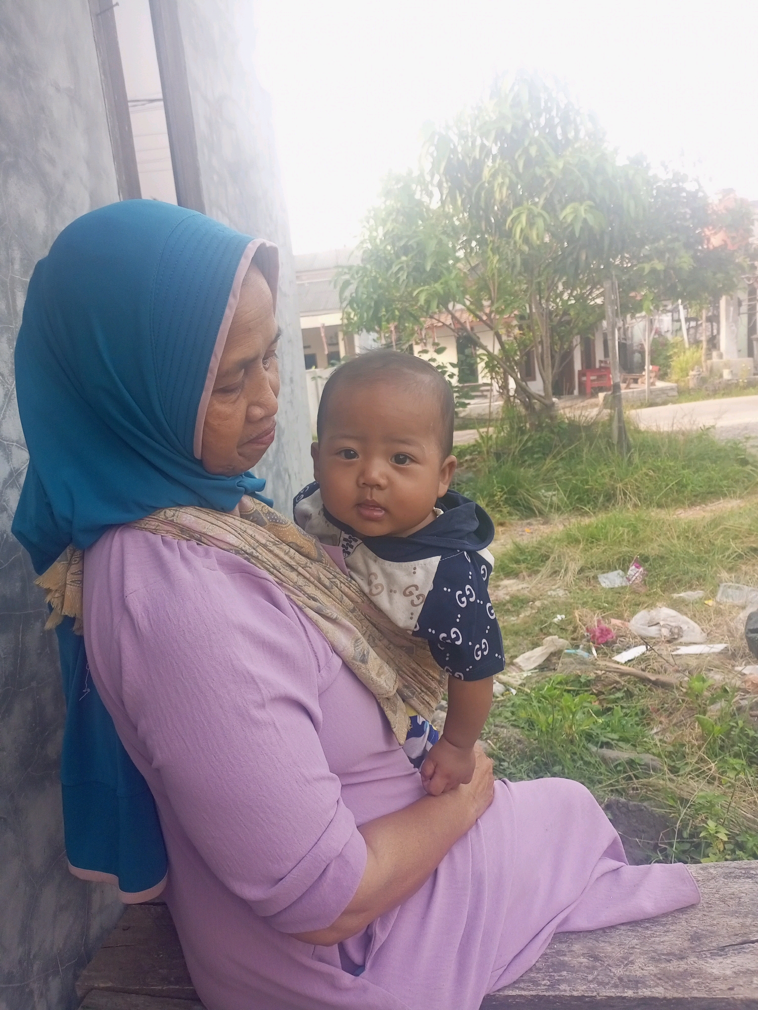 An older woman wearing a blue hijab and a purple dress is sitting on a wooden bench. She is holding a baby, who is wearing a dark blue hooded jacket with a white and gold pattern. The woman is looking down at the baby, and the baby is looking towards the camera. They are sitting outdoors, with a gray wall behind them and a grassy area with some debris in front. In the background, there are trees and houses, suggesting a residential area in the city of Jawilan, Indonesia. The lighting is bright, possibly indicating daylight.
