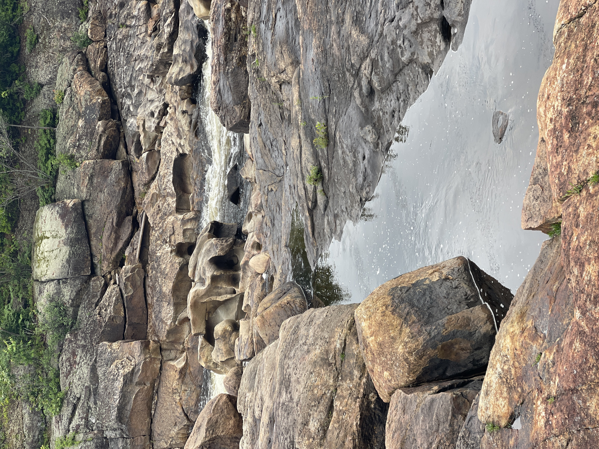 The image depicts a natural rocky landscape with a flowing river. Large, weathered rocks of various brown and gray hues dominate the scene, with some covered in patches of moss and lichen. The river, appearing dark and somewhat turbulent, flows through the rocky terrain, with whitewater visible in some sections. Small green plants and weeds are scattered among the rocks, indicating a natural, possibly wild, environment. The overall impression is one of a rugged, natural setting, likely a riverbed or gorge. There are no people or man-made structures visible in the image.
